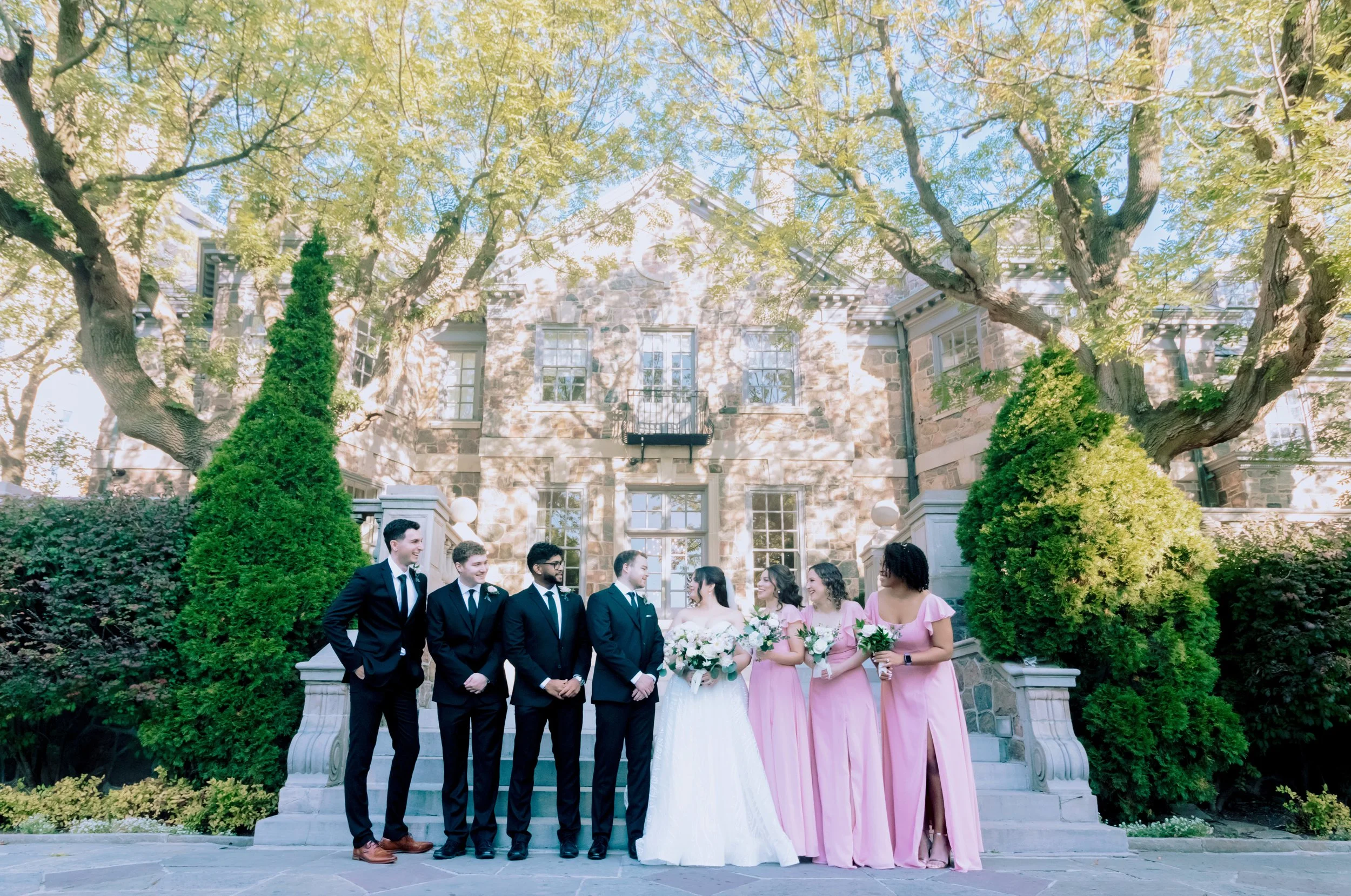 Group of bridesmaids and groomsmen standing outside on steps in front of a stone building with large trees and greenery.