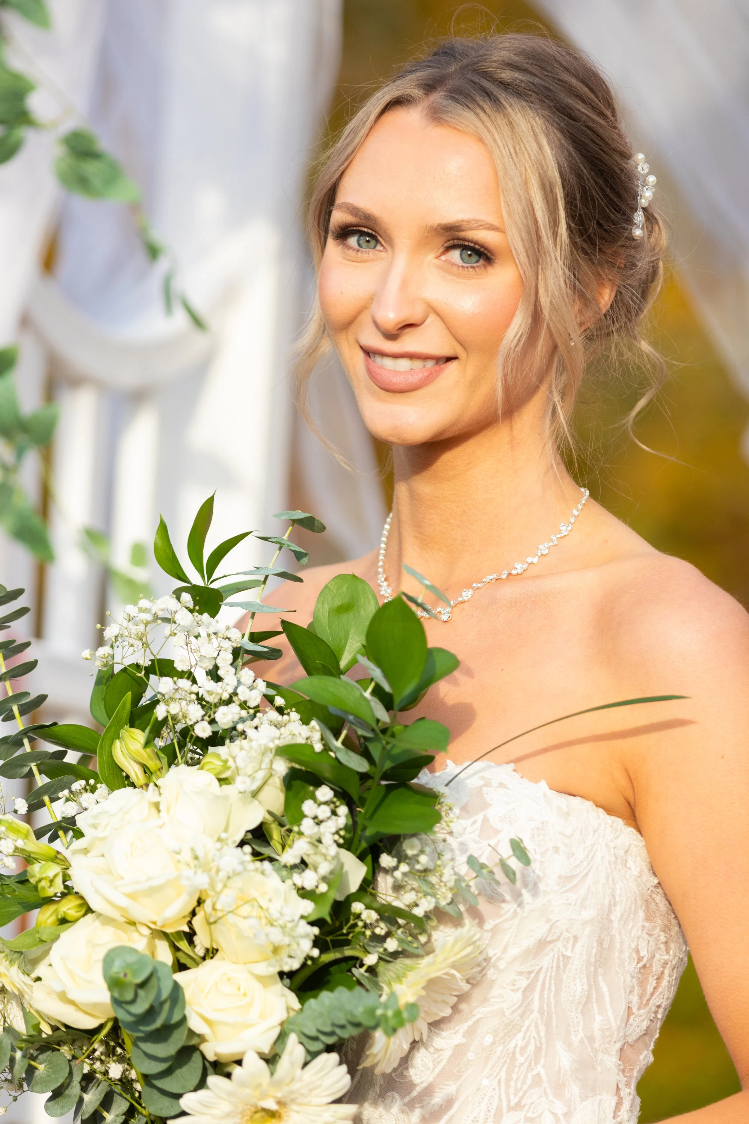 A bride with blonde hair and blue eyes, smiling and holding a bouquet of white flowers and greenery, outdoors during daytime.