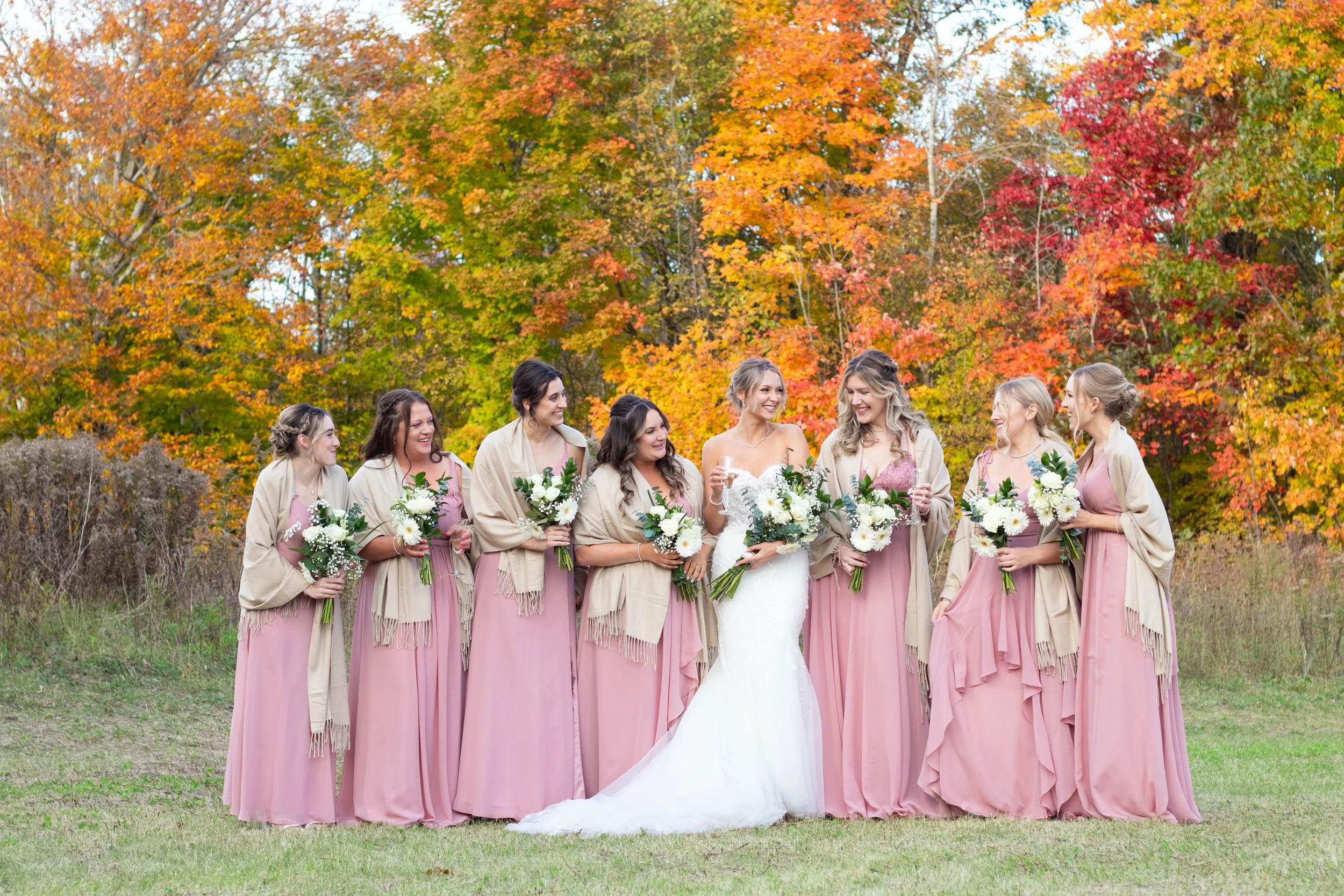 Group of women in pink dresses and beige shawls, holding bouquets, standing outdoors with colorful autumn trees in background.