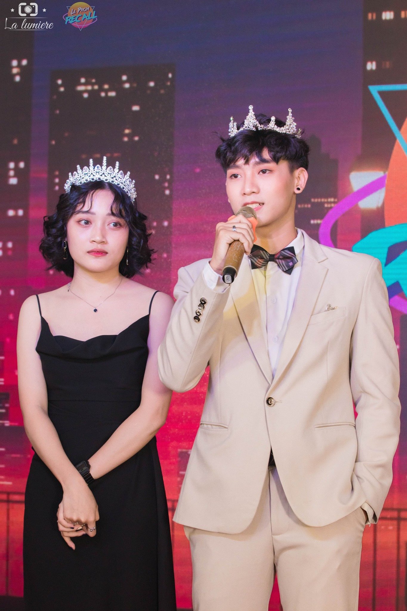 A young man and woman wearing crowns stand on stage, with the man speaking into a microphone, in front of a colorful, cityscape-themed backdrop.