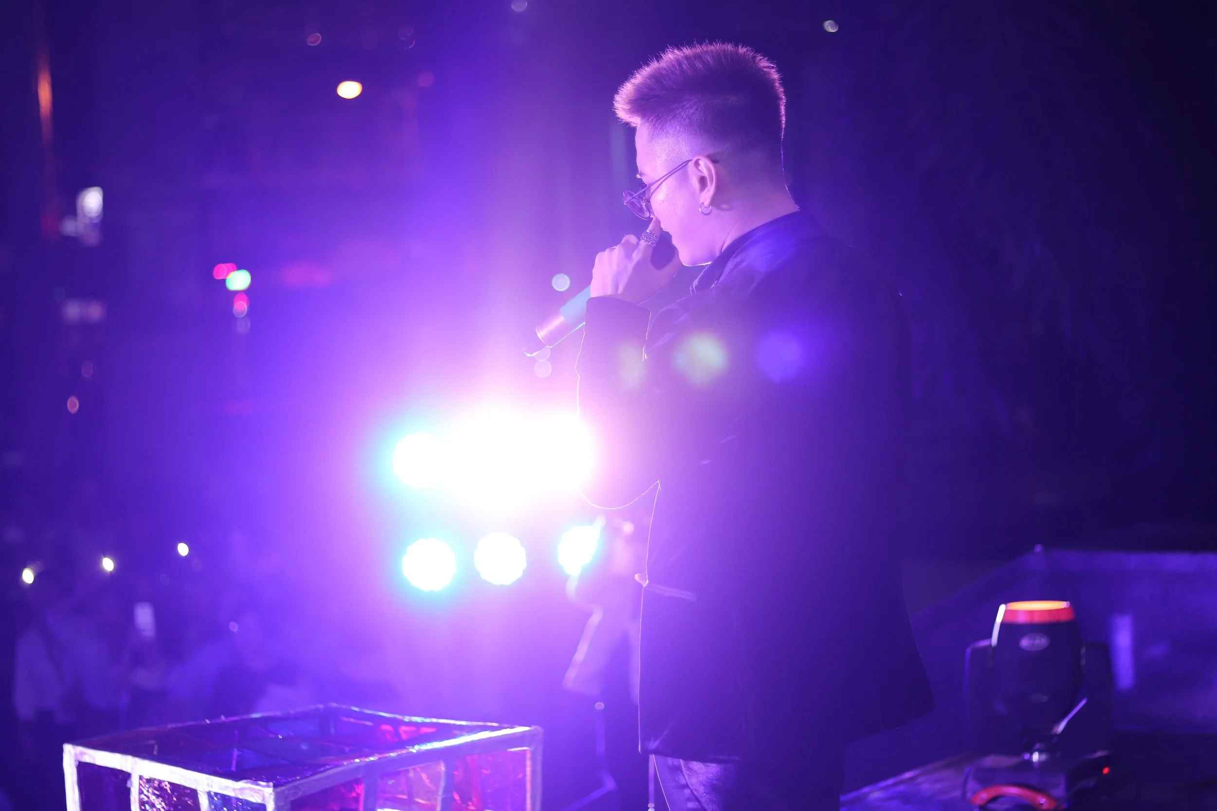 A person with dyed hair and glasses performing on stage at night, holding a microphone with bright purple stage lights around.