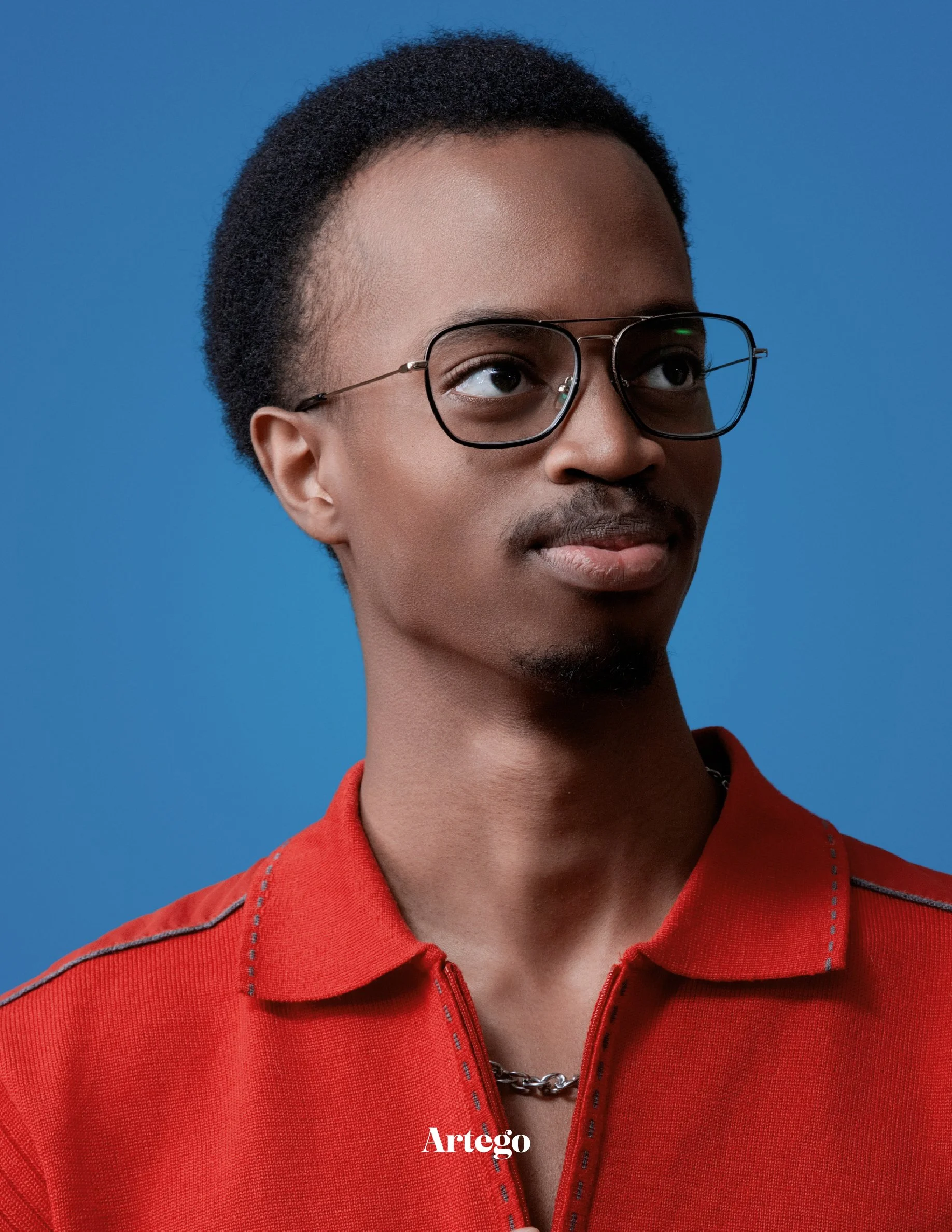 A young Black man with short, natural hair, wearing glasses, a red collared shirt, and a silver chain necklace, looking slightly to the right against a blue background.