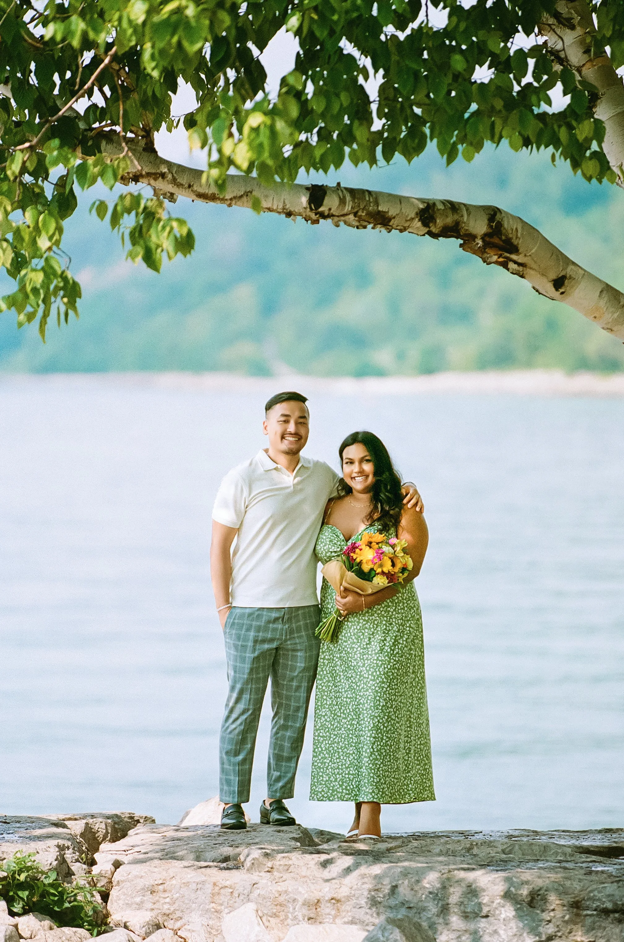 A happy couple standing on rocks near a body of water, smiling, with the woman holding a bouquet of flowers, trees overhead, and a distant green hillside.
