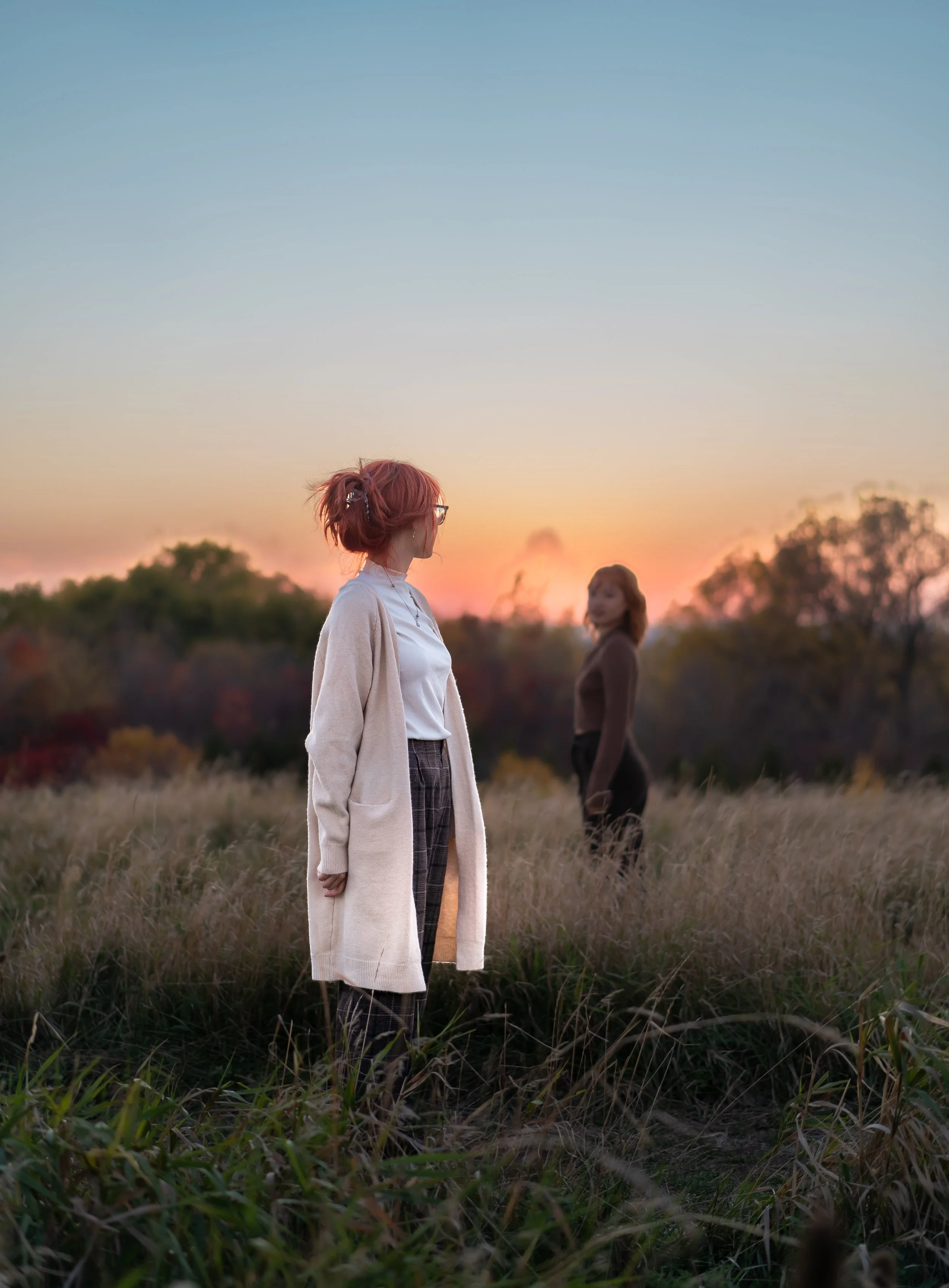 Two women standing in a field at sunset, one in the foreground with pink hair and a beige coat, and the other blurred in the background with brown hair.