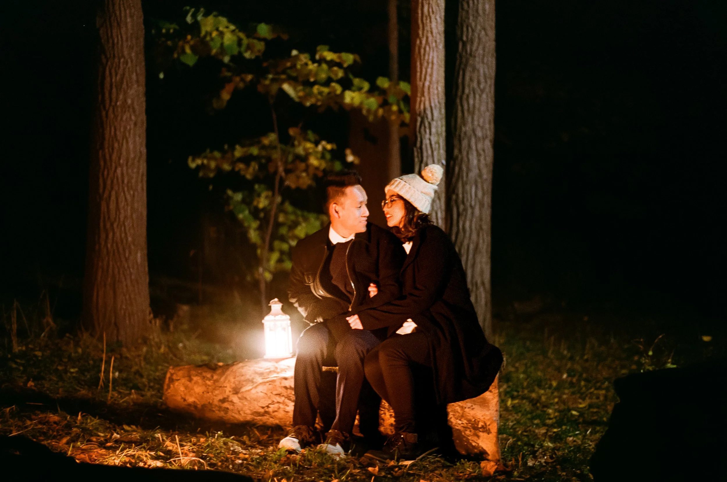 A man and woman sit on a log in a forest at night, illuminated by a lantern, gazing at each other, the woman wearing a beanie and glasses.