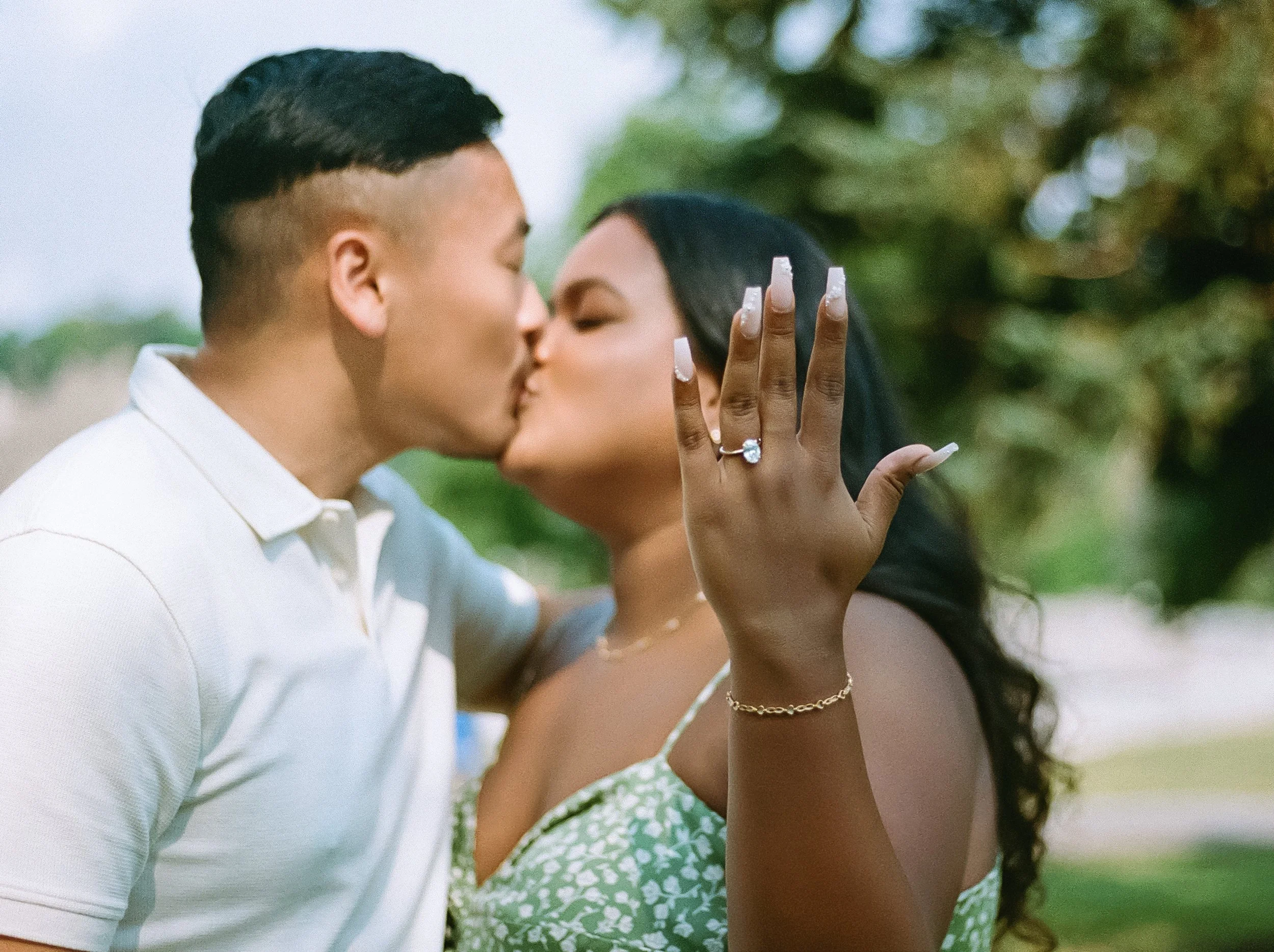 A couple sharing a kiss outdoors, with a woman showing her engagement ring on her raised hand.
