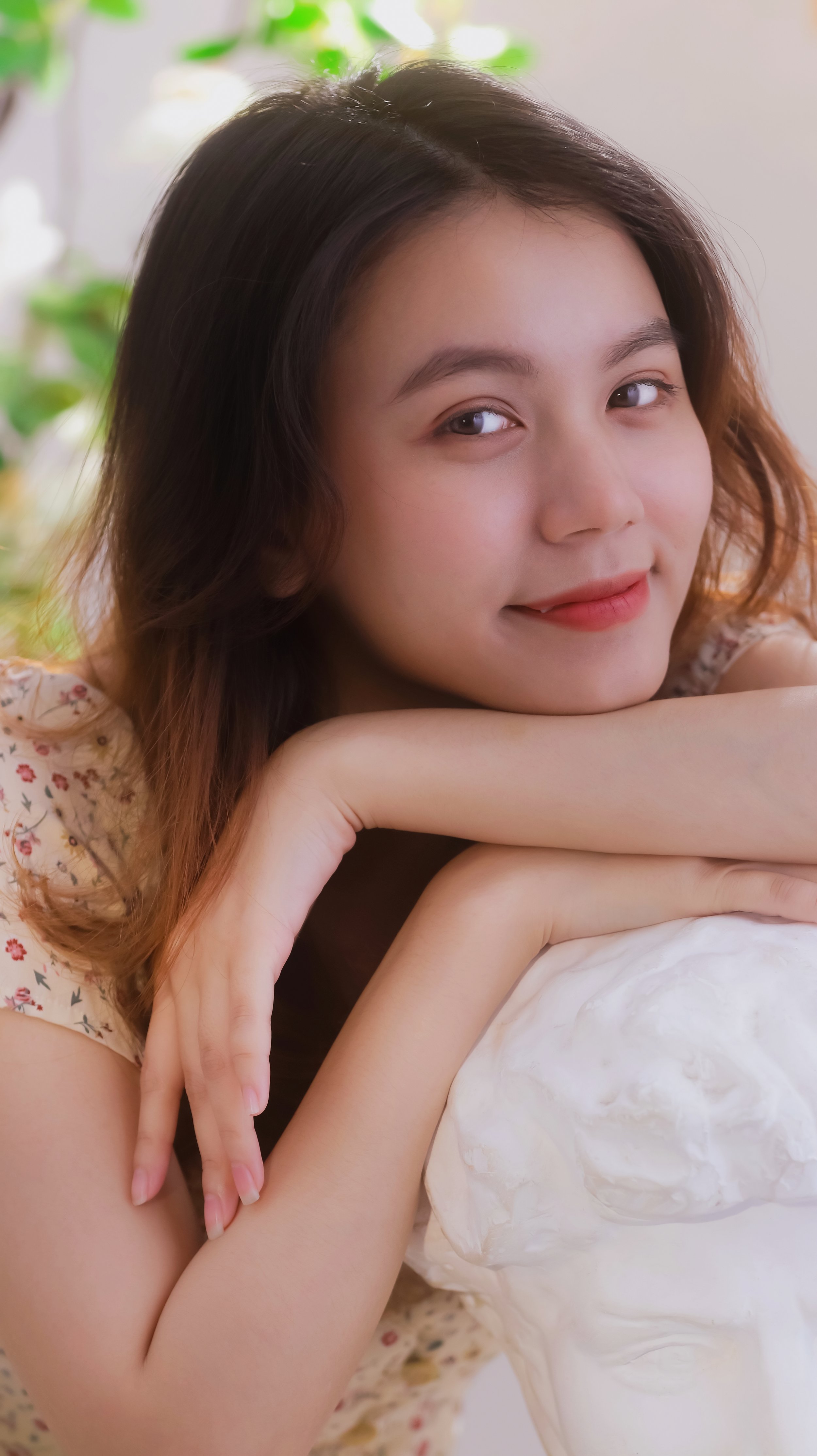 A young woman with wavy brown hair and light skin rests her chin on her arms, leaning on a large white object, possibly a sculpture, with a background of green plants.