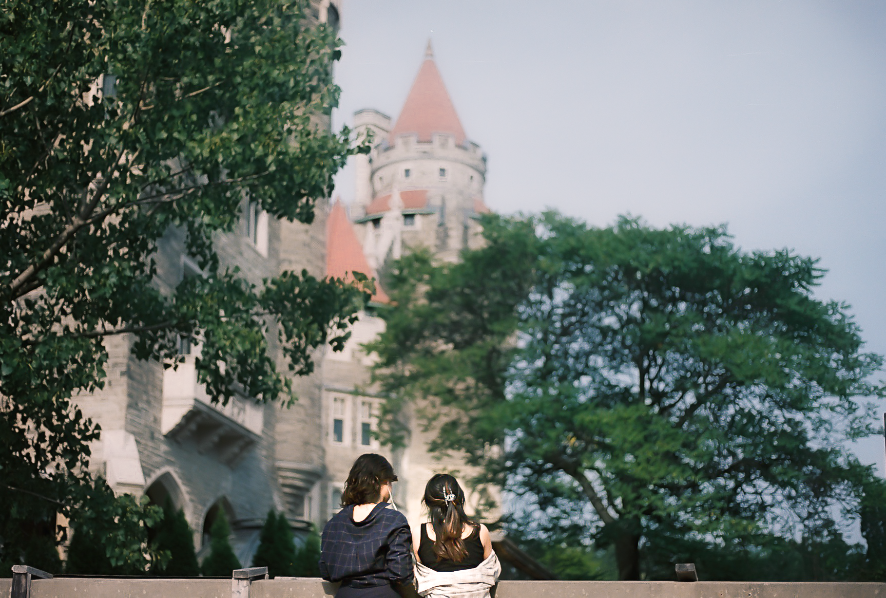 Two people sitting on a bench, facing a castle, trees around, in a park.