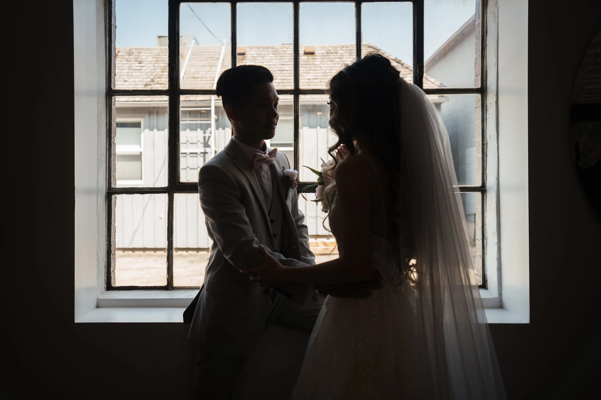 Silhouette of a bride and groom holding hands in front of a window, with outdoor buildings visible outside.