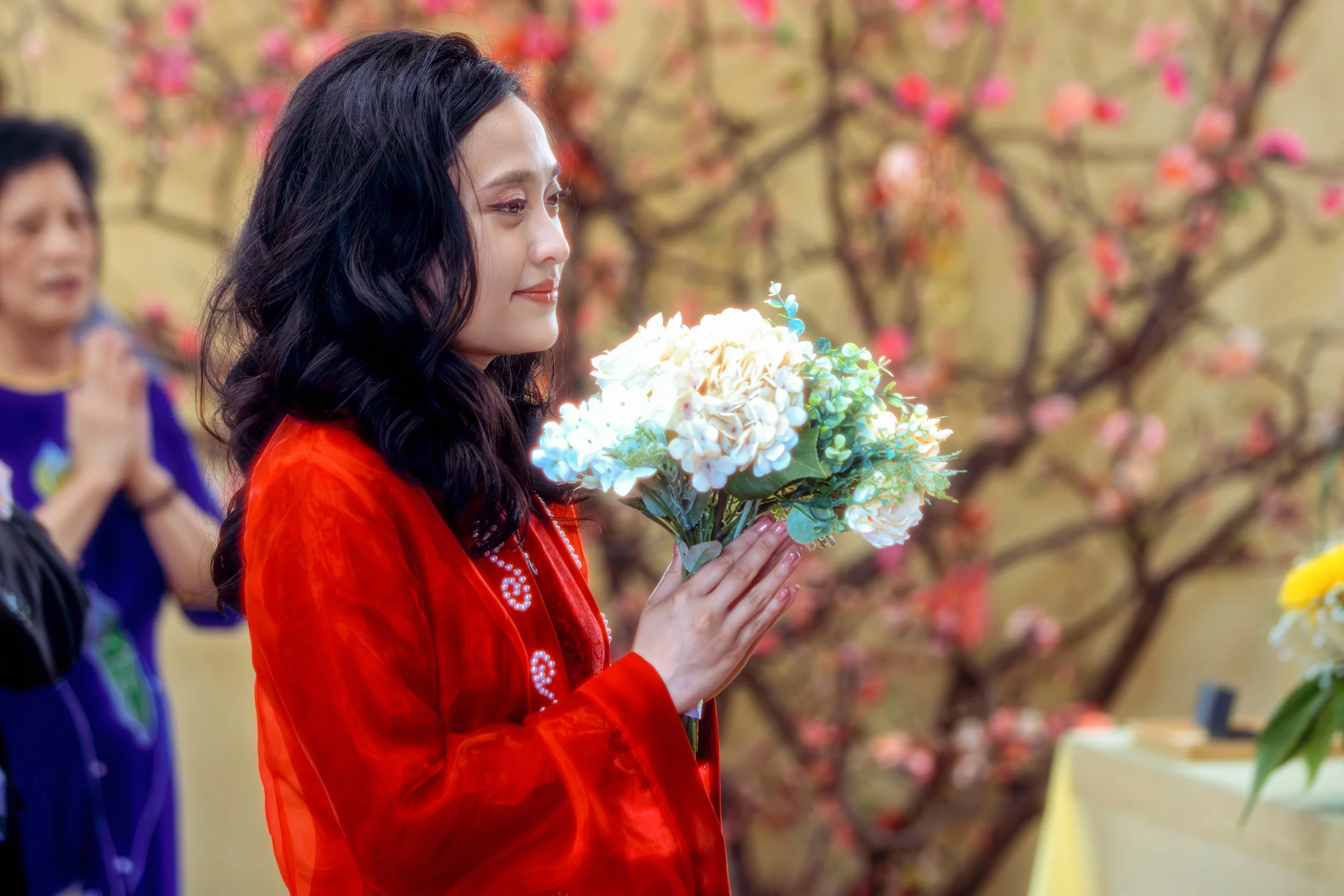 A woman in a red dress holding a bouquet of white flowers, standing in front of a background of pink flowering trees.