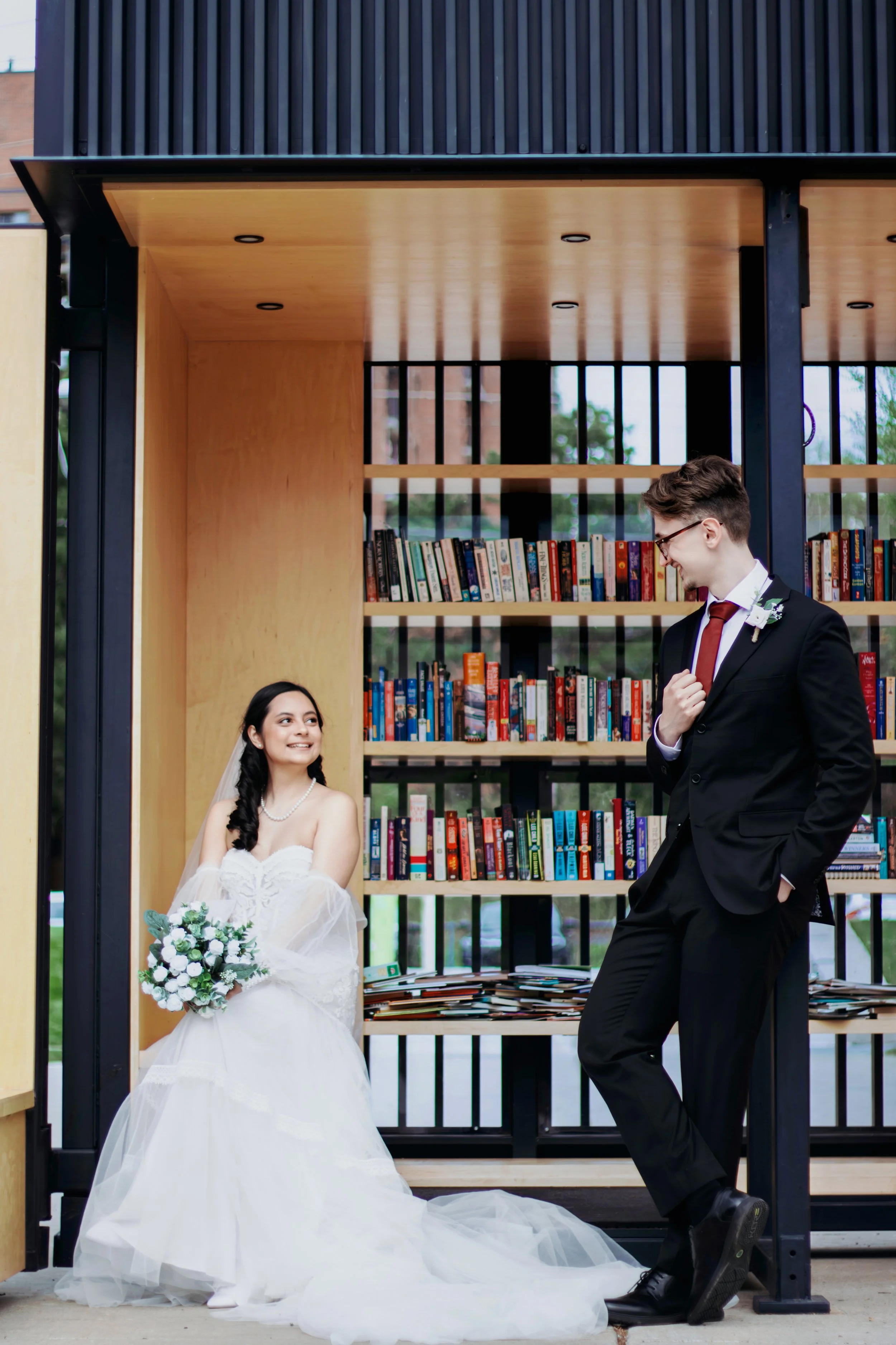 Bride in a white wedding dress and veil holding a bouquet of white flowers smiling at groom in a black suit and red tie outside near a bookshelf.