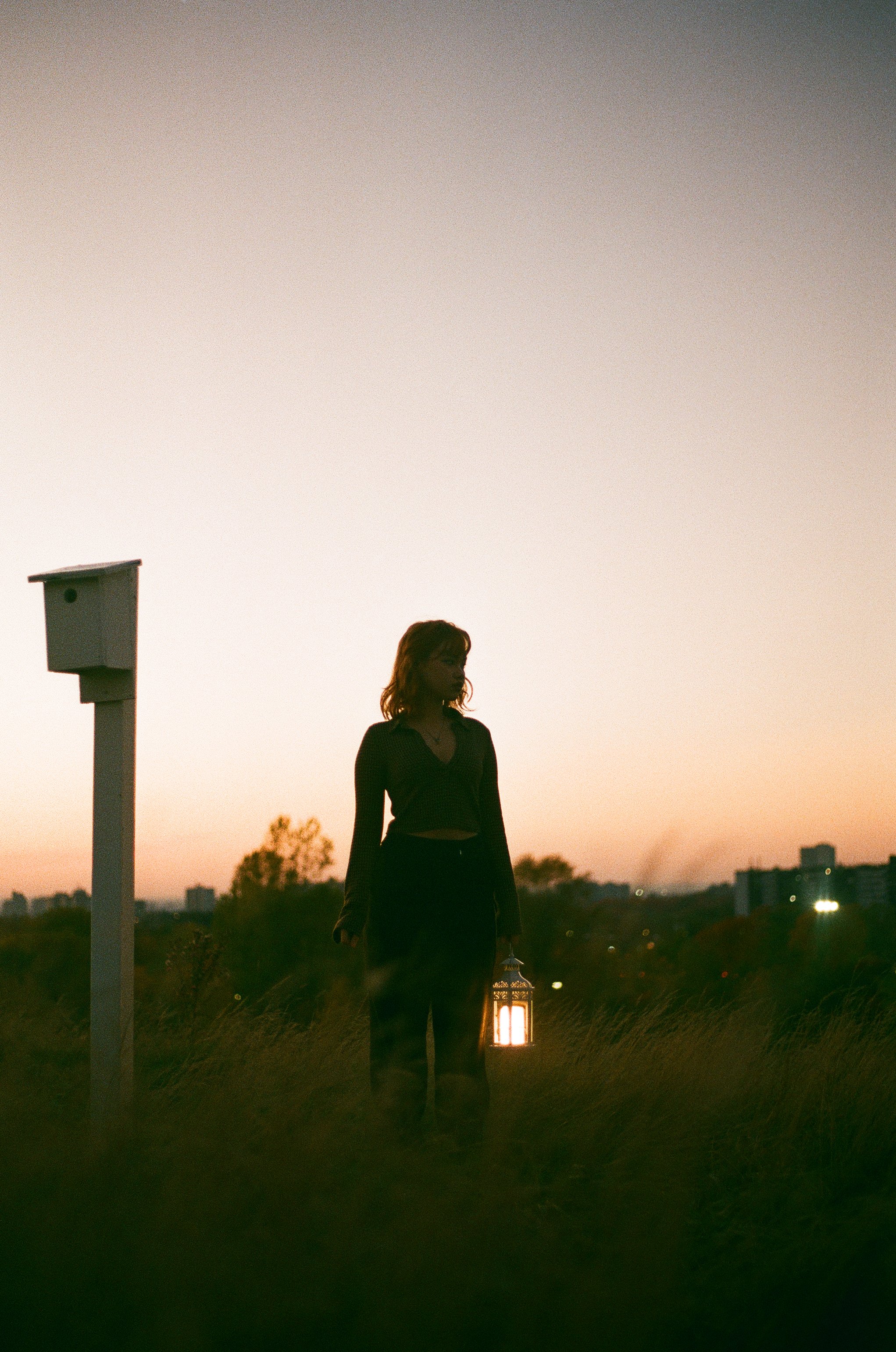 A woman holding a lantern standing outdoors during sunset with a cityscape in the background.