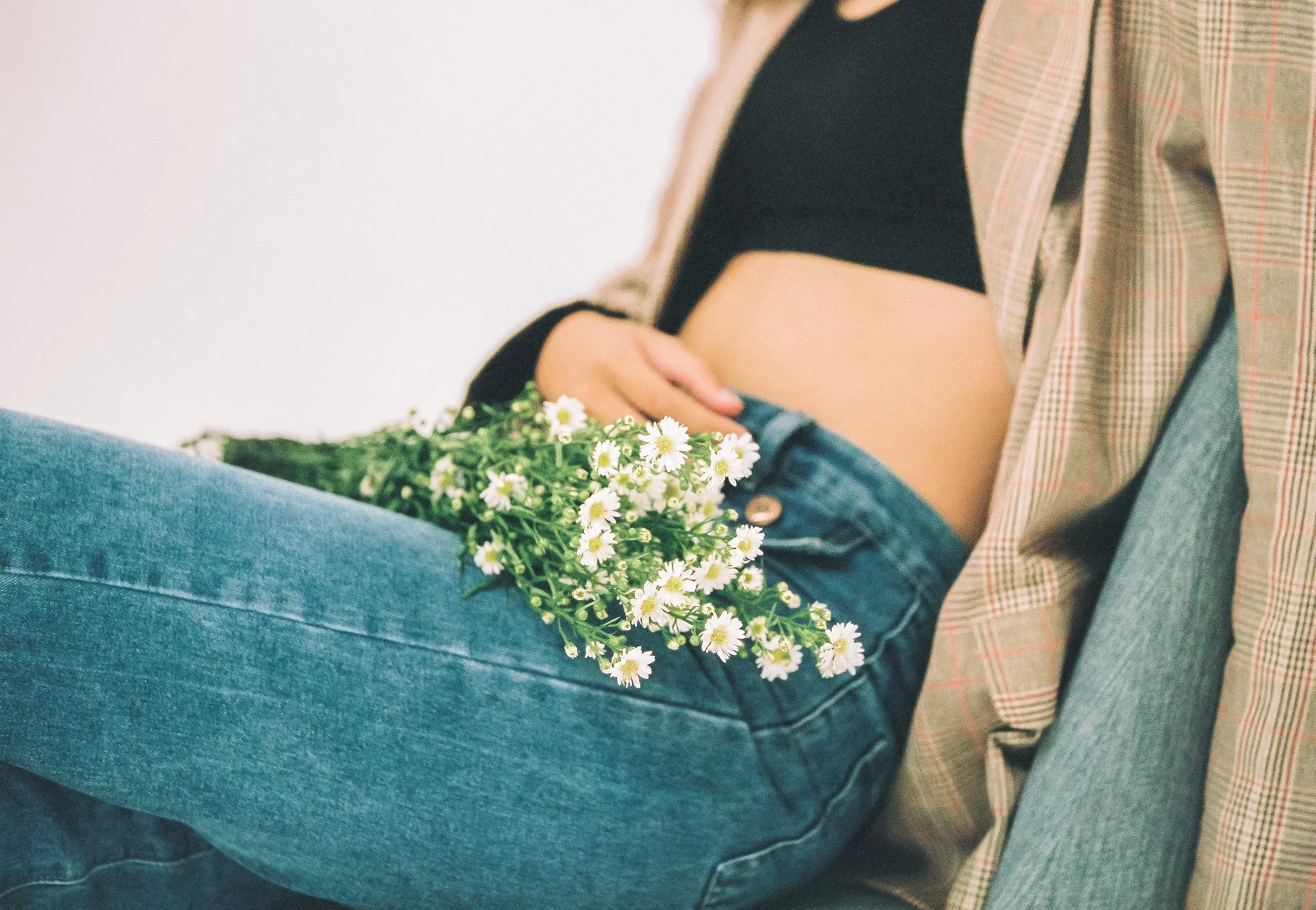 A pregnant woman sitting on a chair, holding a bunch of white daisies with yellow centers in her lap. She is wearing a black crop top, blue jeans, and an open plaid shirt.