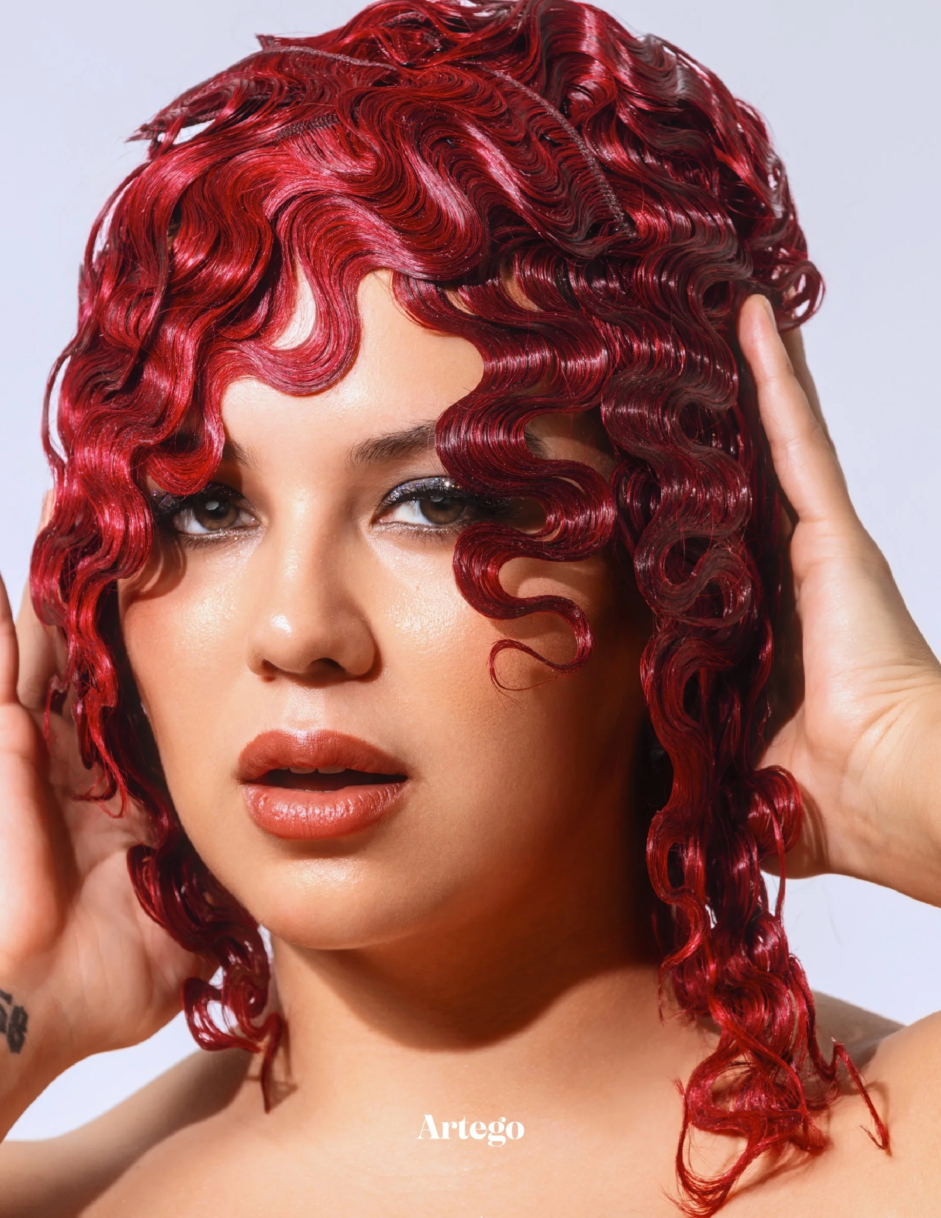 Close-up of a woman with curly red hair, touching her hair with both hands, and looking into the camera.