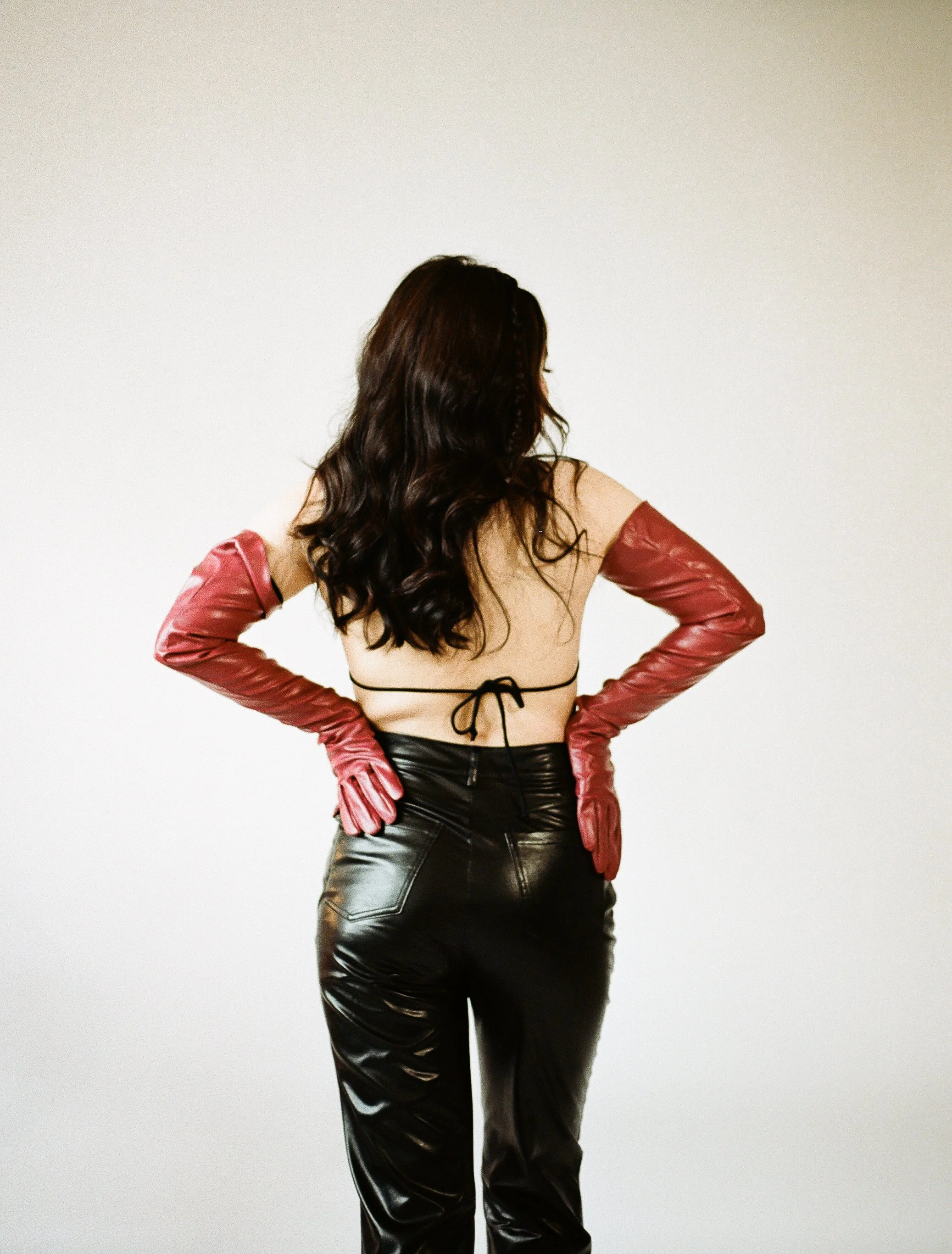 A woman with long dark wavy hair wearing black shiny leather pants, a bare back top, red leather gloves, standing against a plain white background.