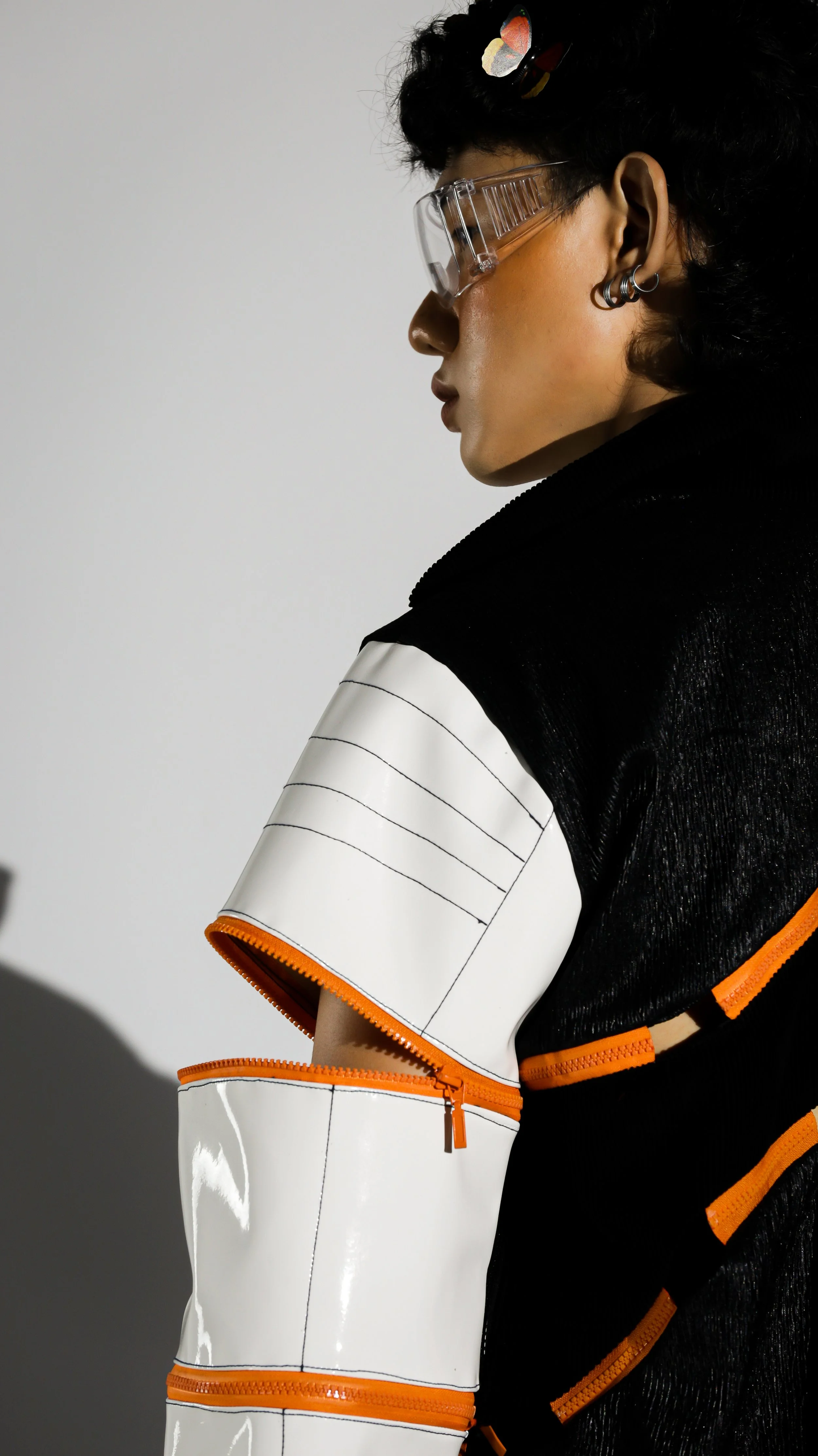 Side profile of a person with short black curly hair, wearing clear safety glasses, multiple earrings, and fashion-forward clothing with black, white, and orange accents, against a plain background.