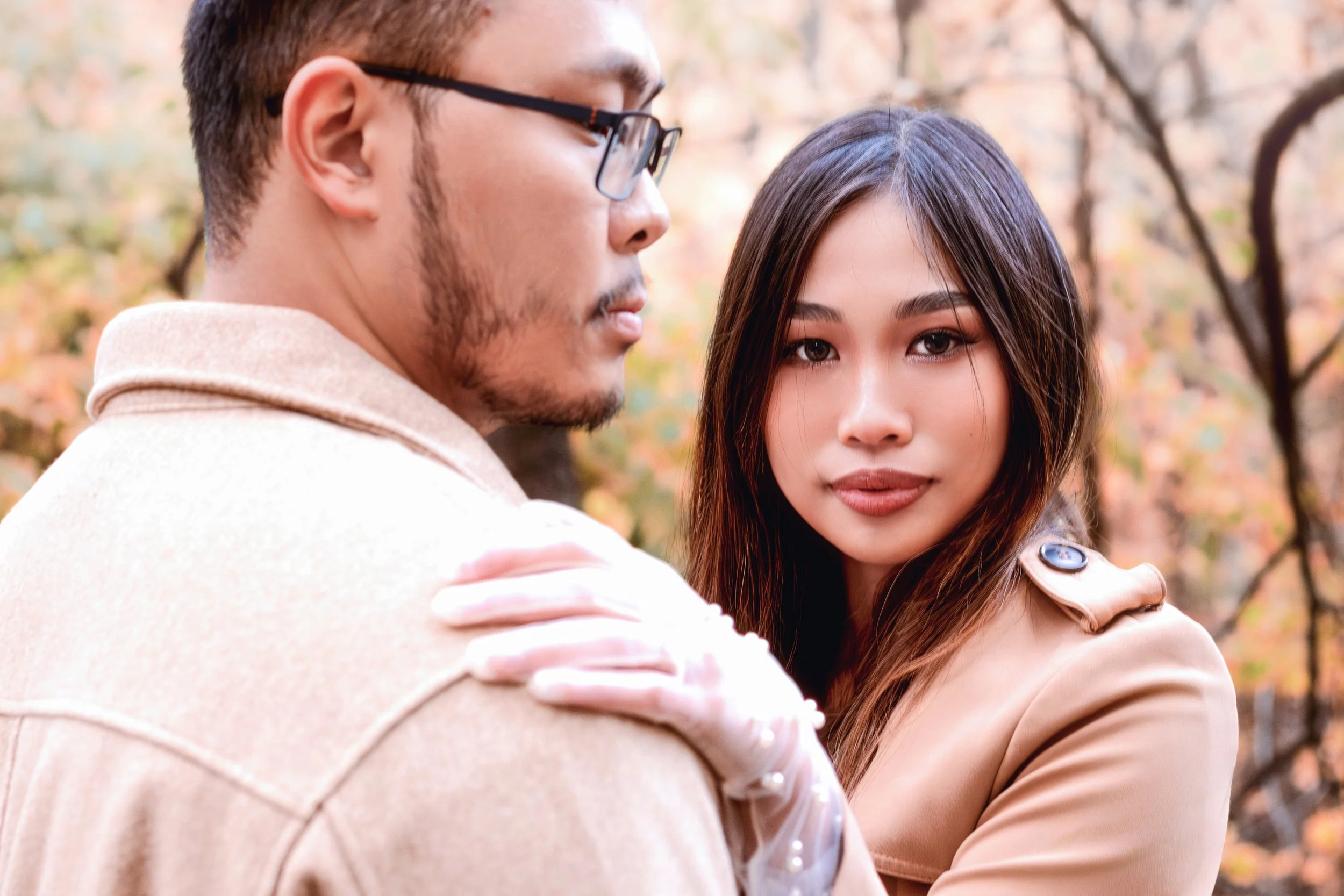 A man and woman in an outdoor setting with fall foliage, embracing each other while looking at the camera. The woman has long dark hair and the man is wearing glasses.