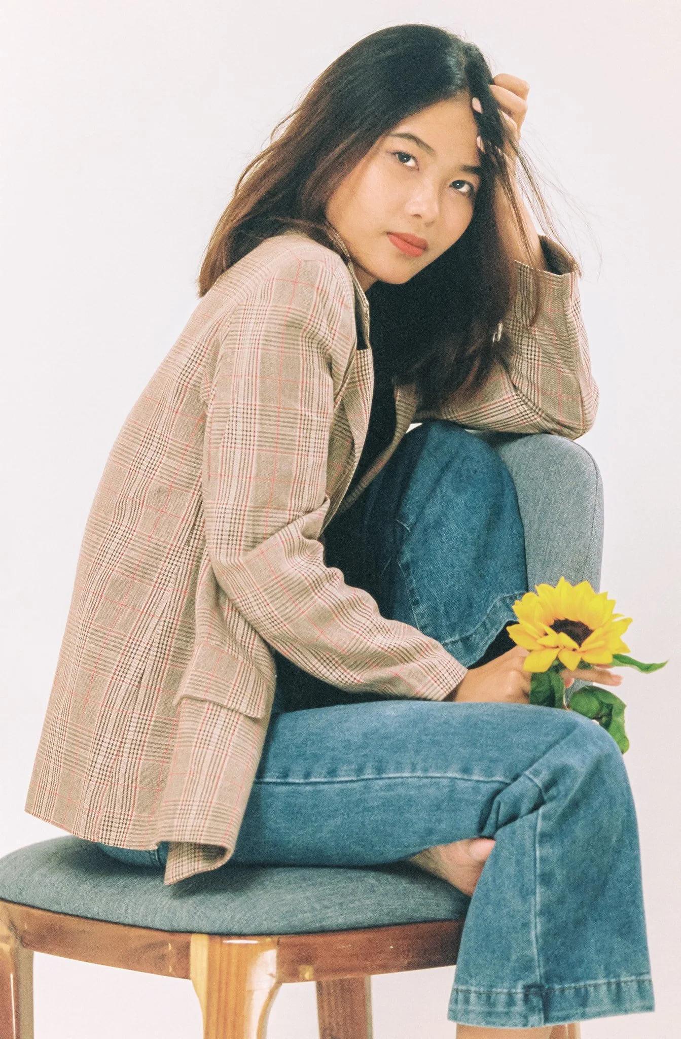 A woman with dark hair sits on a wooden chair, holding a sunflower. She wears a plaid blazer and jeans, with her hand in her hair and a neutral expression.