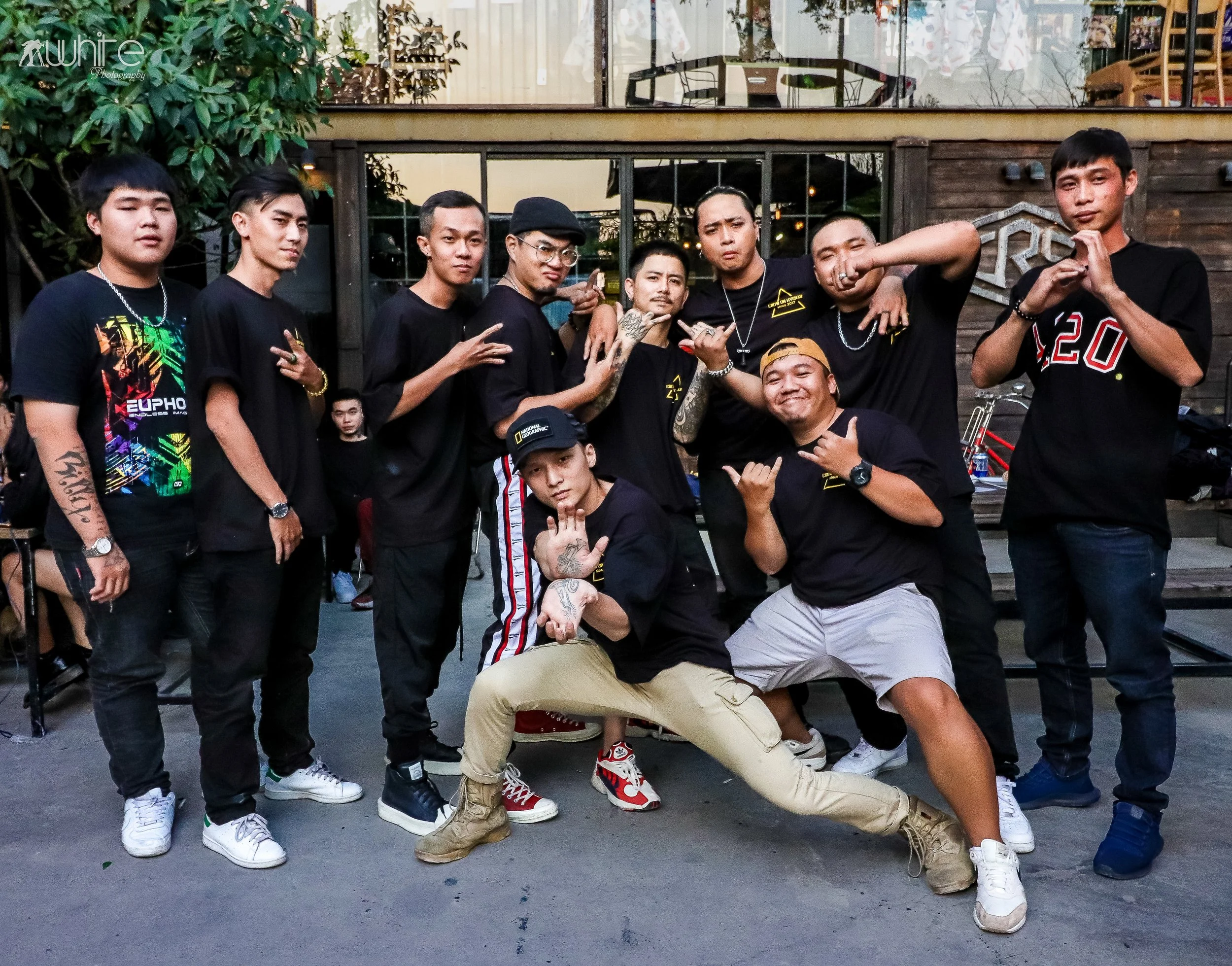 A group of young men posing outdoors in front of a building with large windows, some making hand gestures or flexing their muscles, dressed in casual clothes including black t-shirts, jeans, and sneakers.