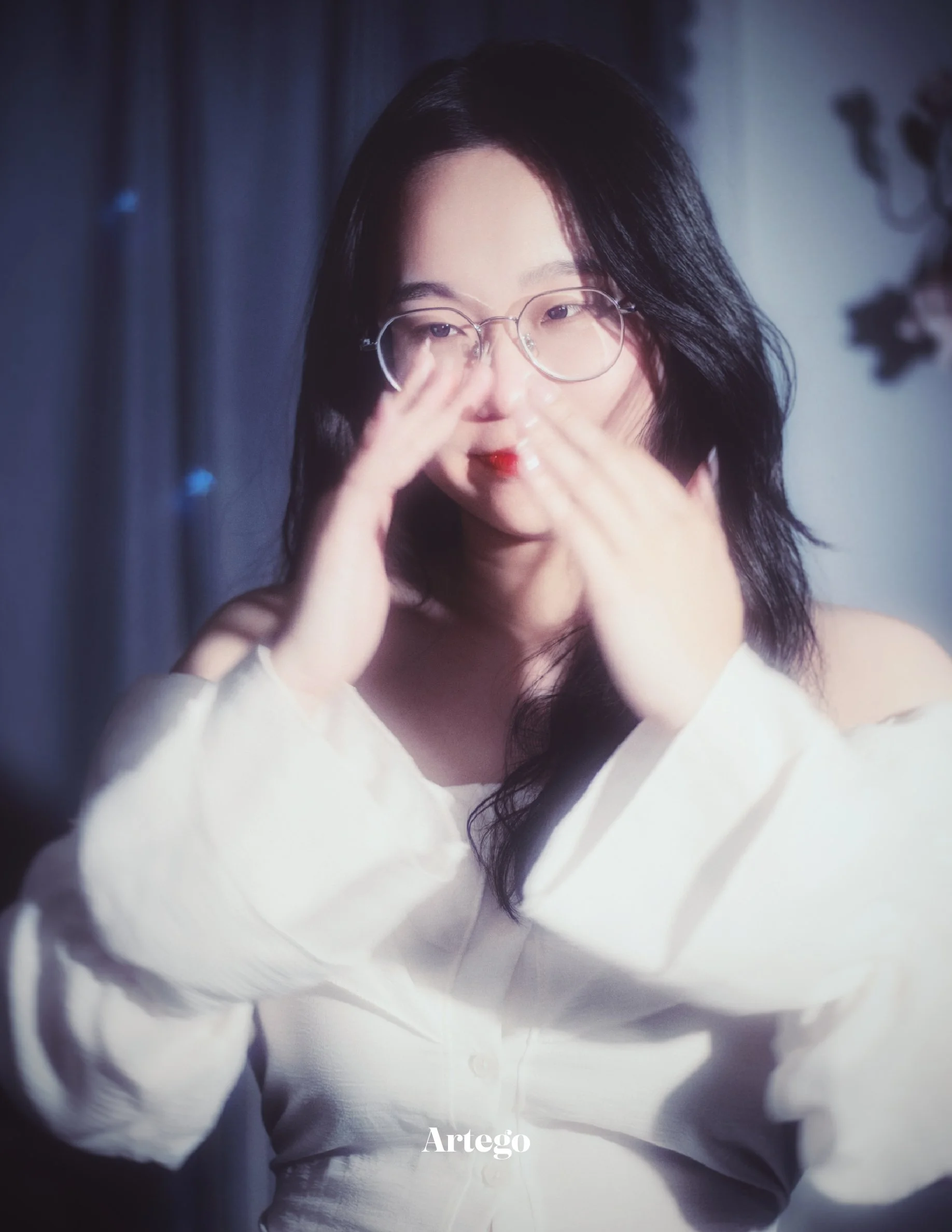 A woman with long black hair, glasses, and wearing a white off-the-shoulder top is touching her face with both hands.