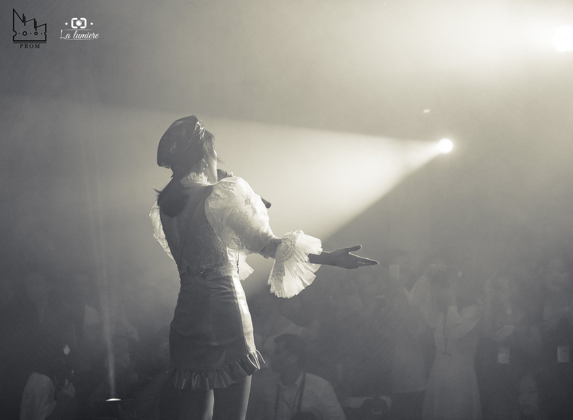 A woman singing on stage, illuminated by bright stage lights, with an audience in the background.