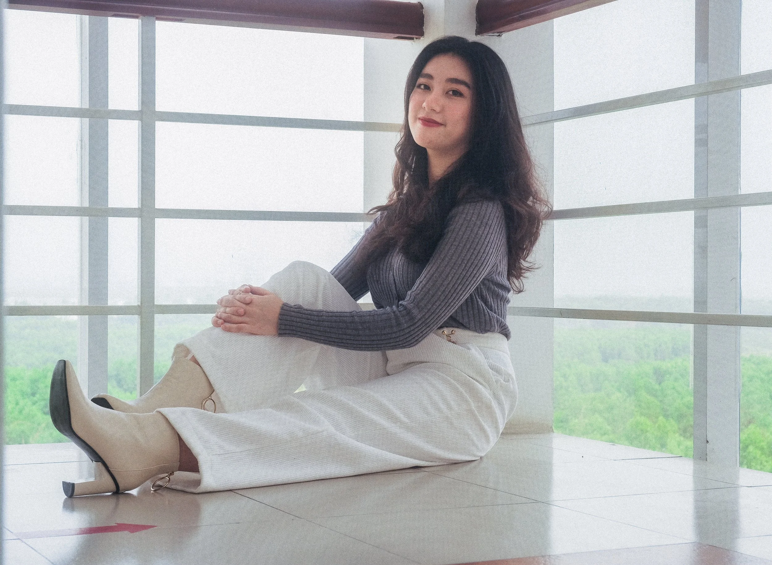 A young woman with dark hair sitting on the floor near large windows, wearing a gray long-sleeve top, white pants, and beige high-heeled boots, with a view of green trees outside.