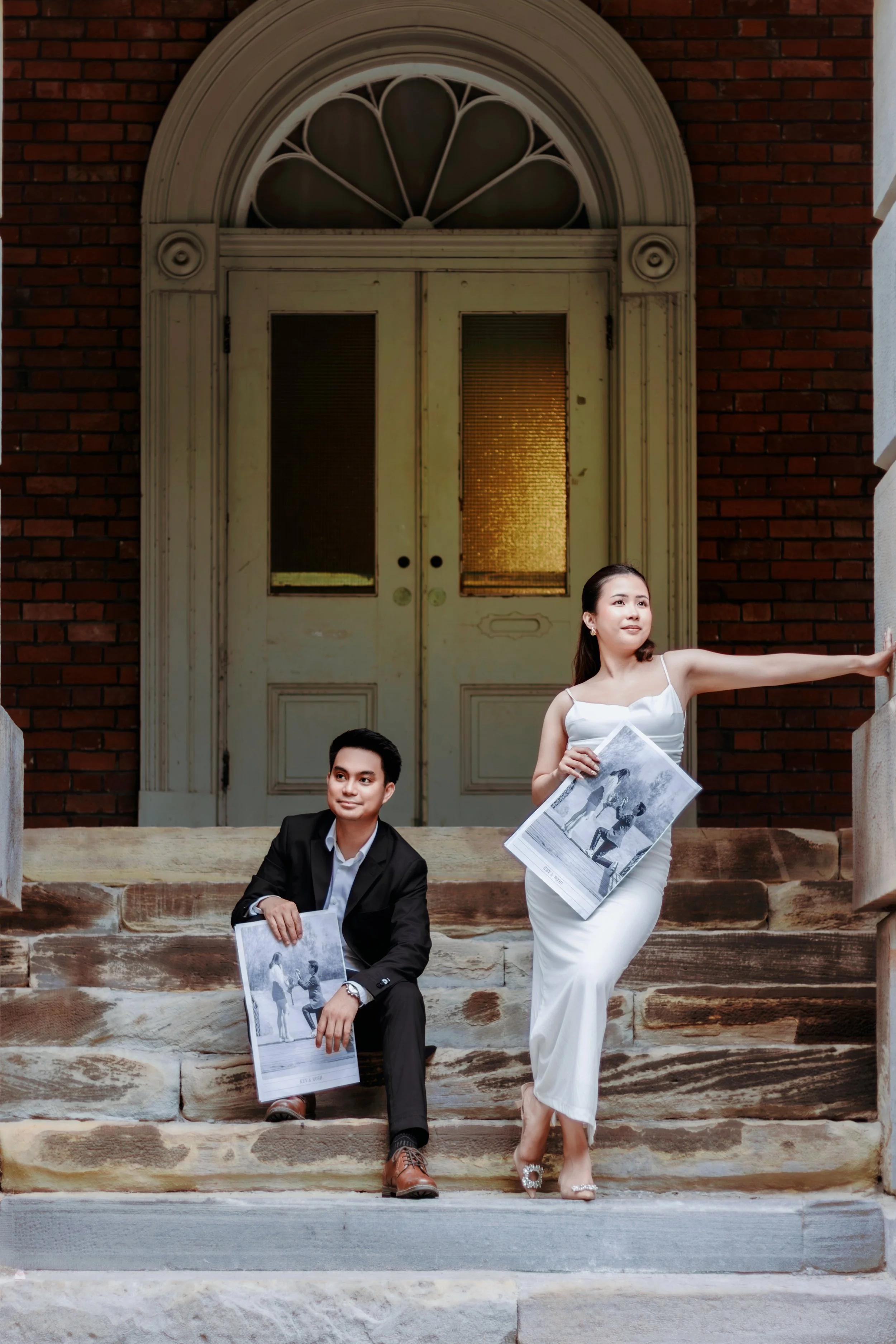 A man in a black suit and a woman in a white dress sitting on stone steps in front of a vintage door, holding newspapers with black-and-white images, on a brick building entrance.