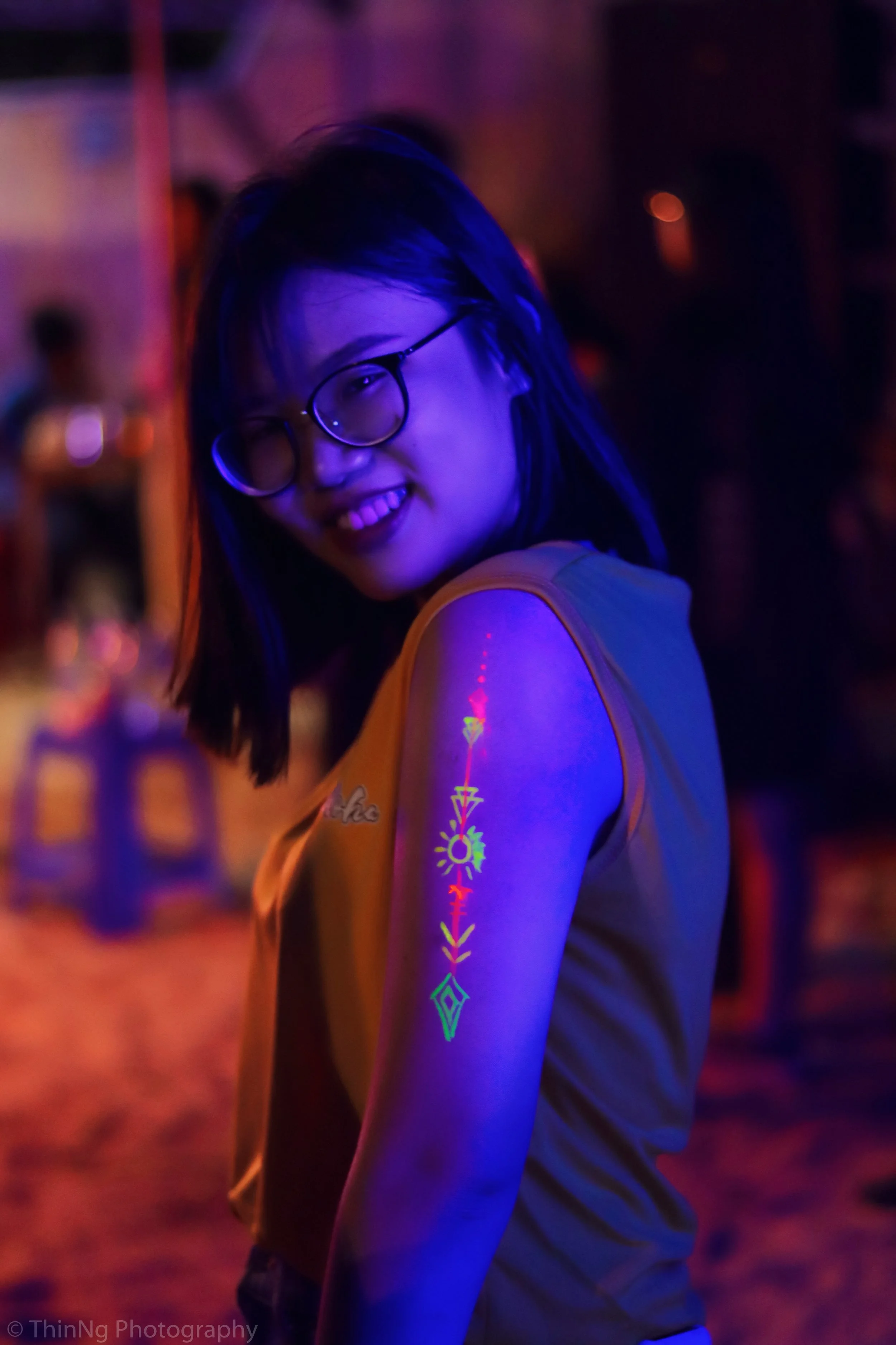 A young woman with glasses, smiling and looking at the camera, is illuminated with blacklight. She is showing a glow-in-the-dark tattoo on her arm, with neon colors and geometric patterns. The background is dark and blurry.
