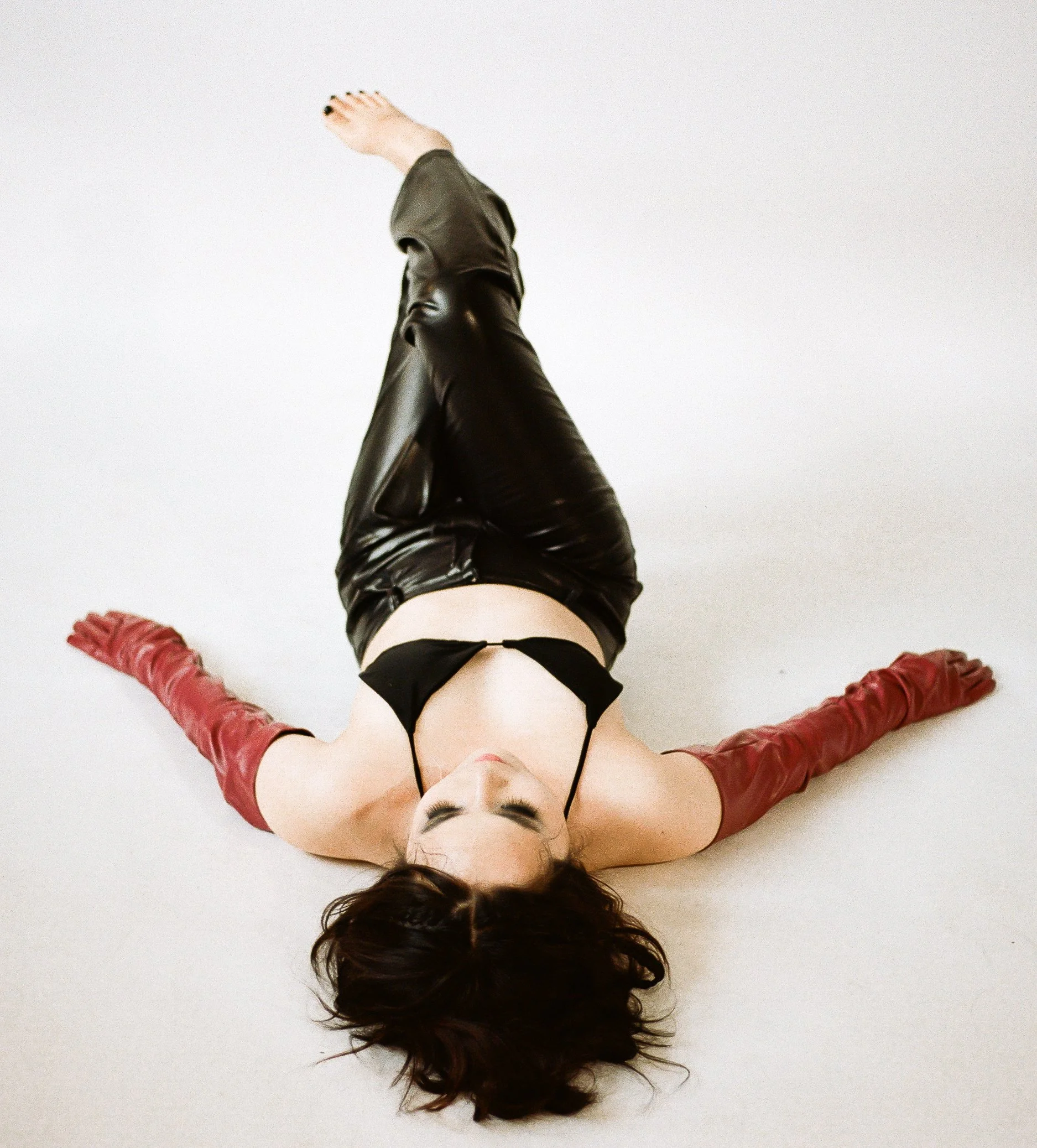 A woman lying on her back on a white surface with her arms spread out, wearing red gloves, black bra, and black leather pants.