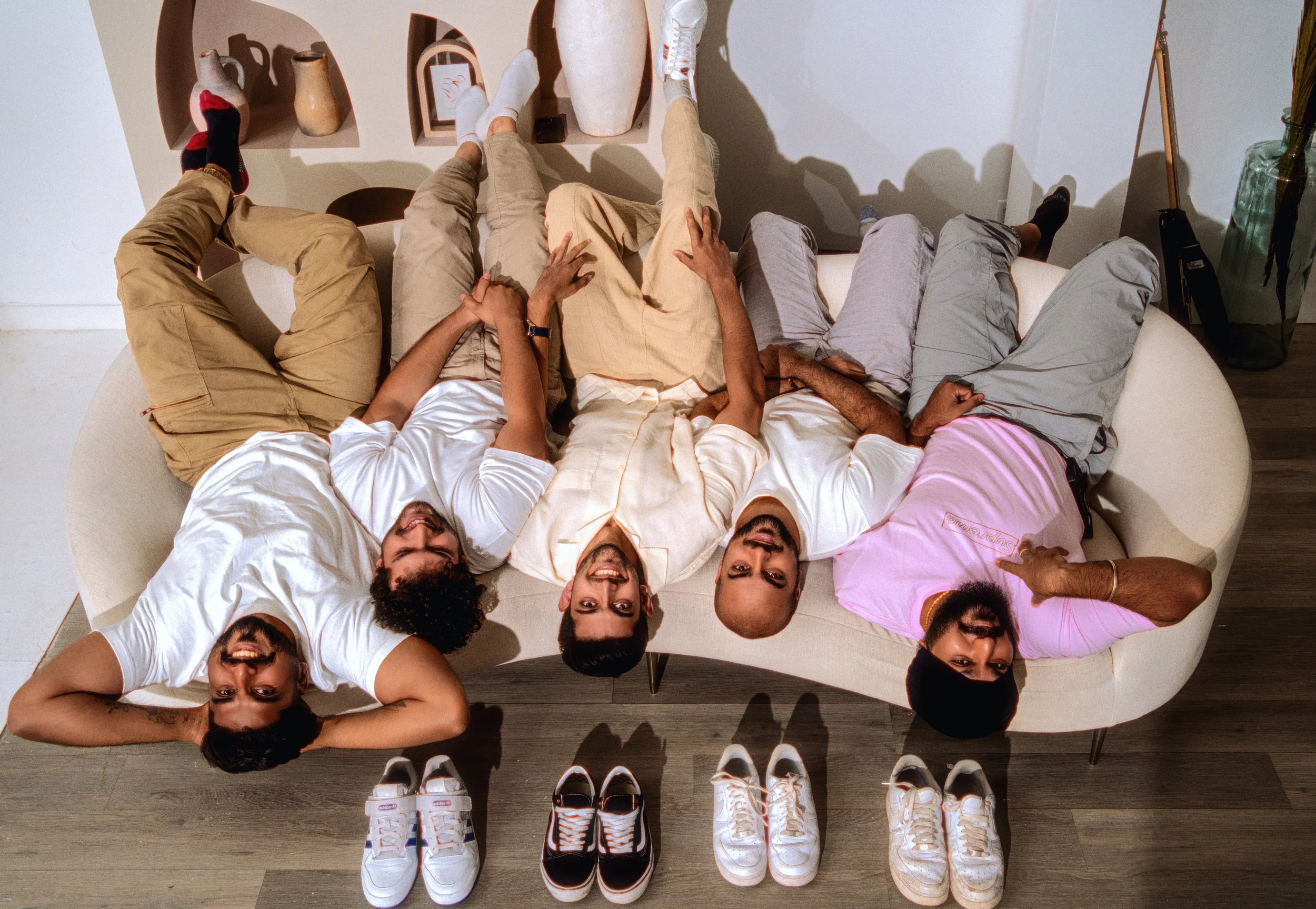 Five people lying upside down on a sofa with their heads at the bottom and feet at the top of the image, smiling and laughing. They are in a living room with three pairs of shoes placed on the floor in front of the sofa.