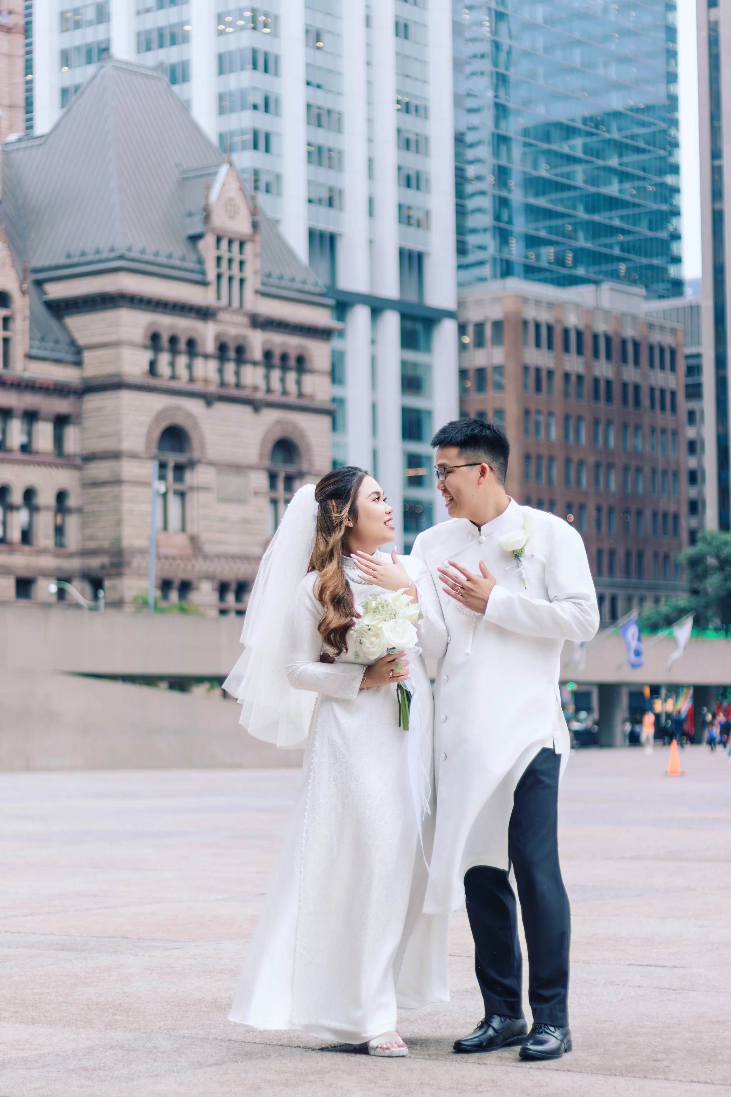 A bride and groom in traditional wedding attire share an intimate moment outdoors in an urban cityscape with tall buildings and historic architecture in the background.