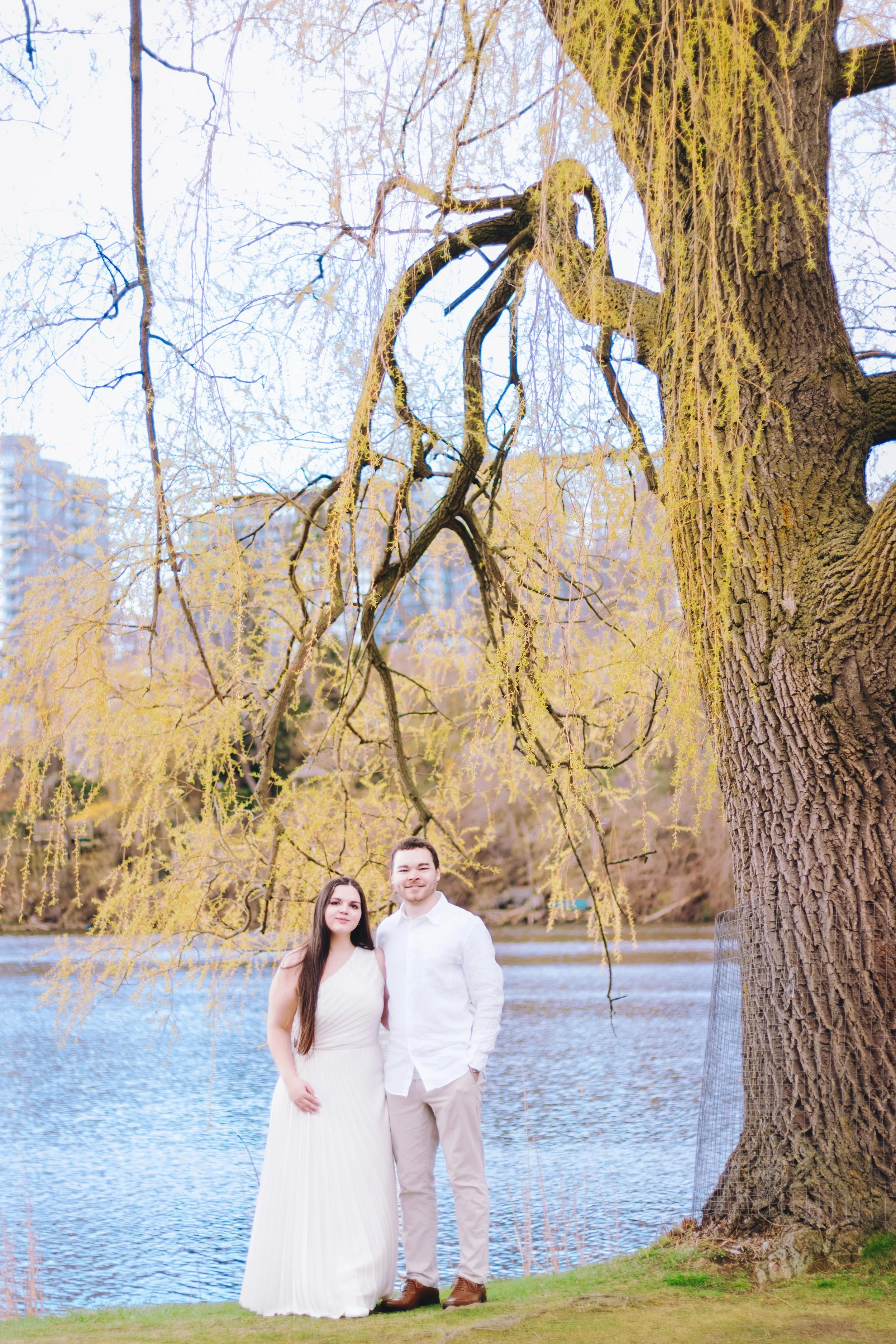 A young woman in a white dress and a young man in a white shirt and beige pants standing by a lake with a large tree with yellowish-green leaves overhead.