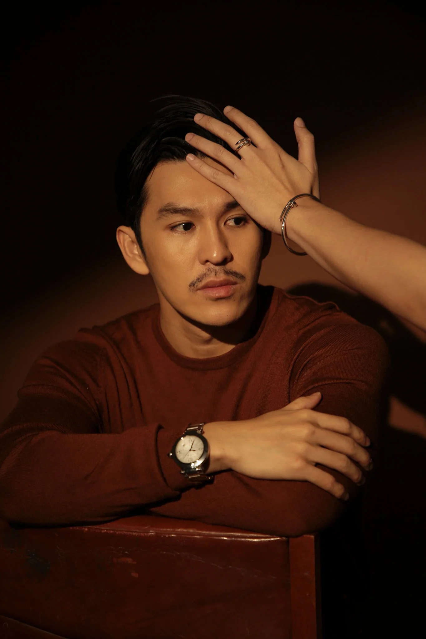 A man with dark hair and a mustache wearing a brown long-sleeve shirt, accessorized with a watch and bracelets, posed with one hand on his forehead and the other resting on a wooden surface against a dark background.