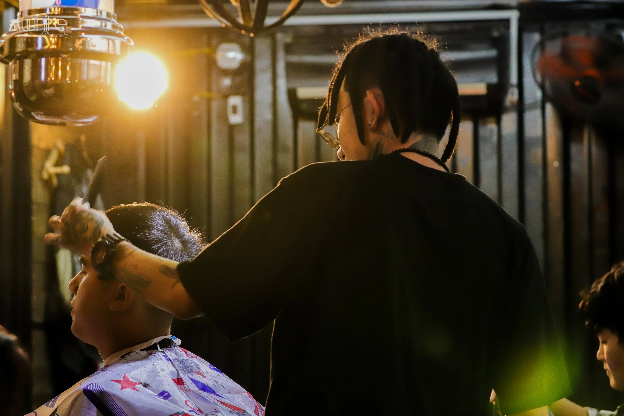 A barber with tattoos and dreadlocks cuts a young man's hair outside during sunset.