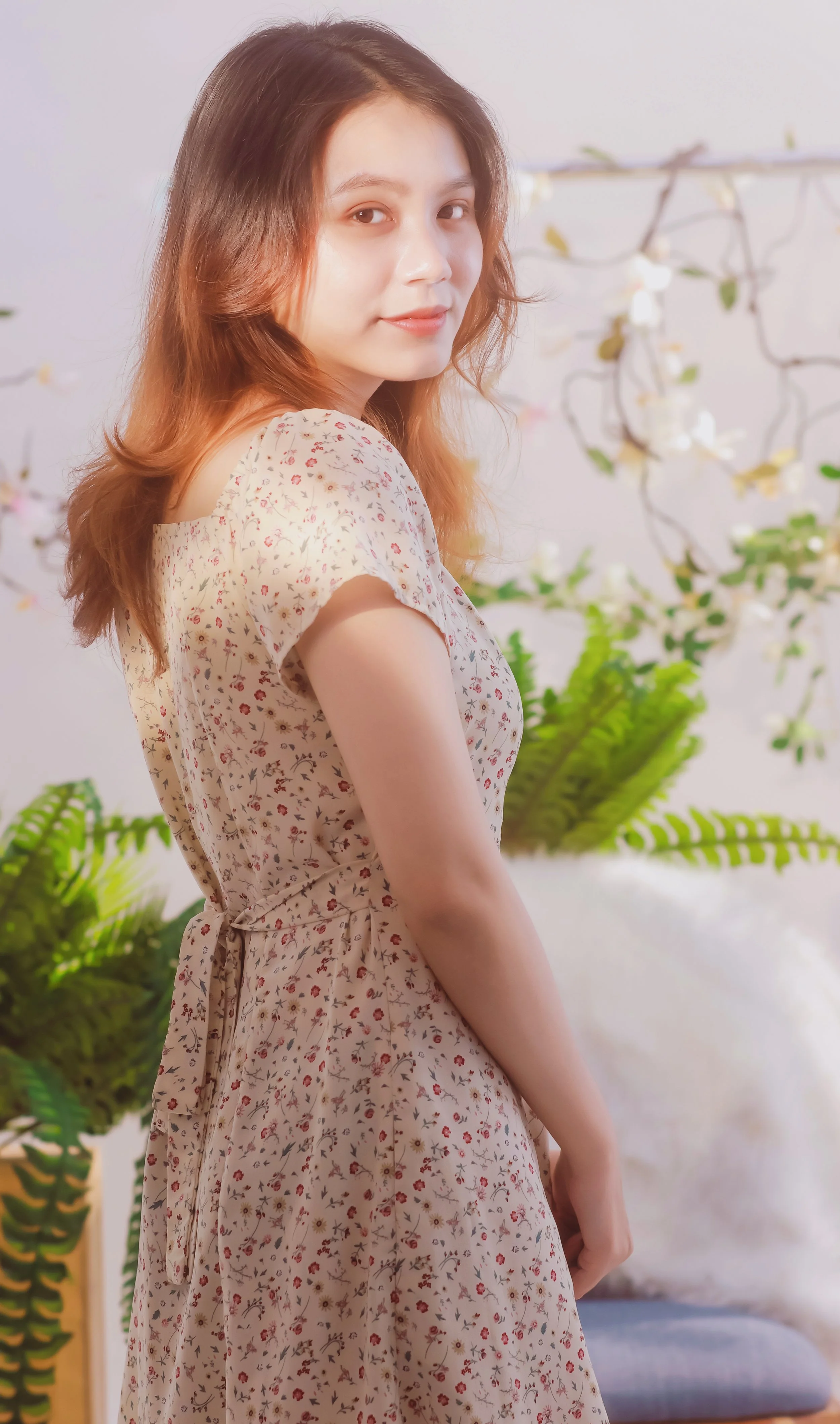 Portrait of a young woman with wavy reddish-brown hair wearing a floral dress, standing indoors with plants and a white wall in the background.