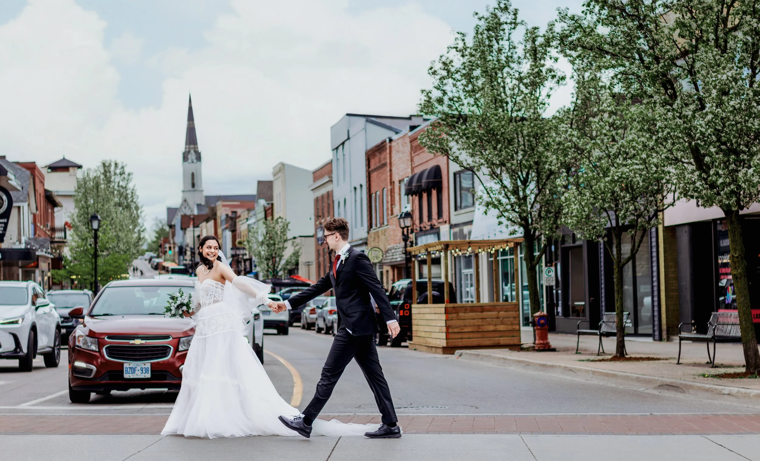 A newlywed couple walking hand-in-hand on a city street, with the bride in a white wedding gown holding a bouquet and smiling, and the groom in a black suit and tie. They are crossing the street with parked cars, storefronts, trees, and a church stee