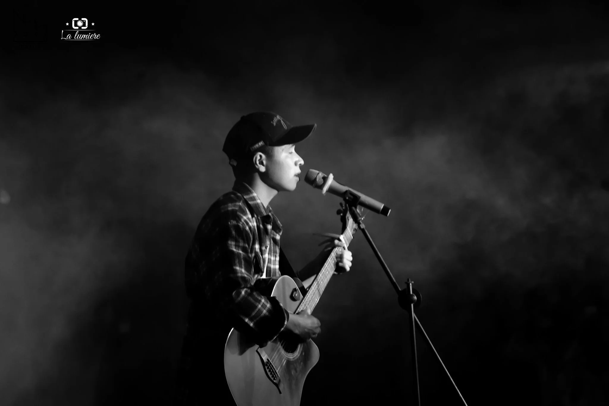A young person wearing a hat and plaid shirt performs music on stage with a guitar and microphone, with a dark background and atmospheric lighting.