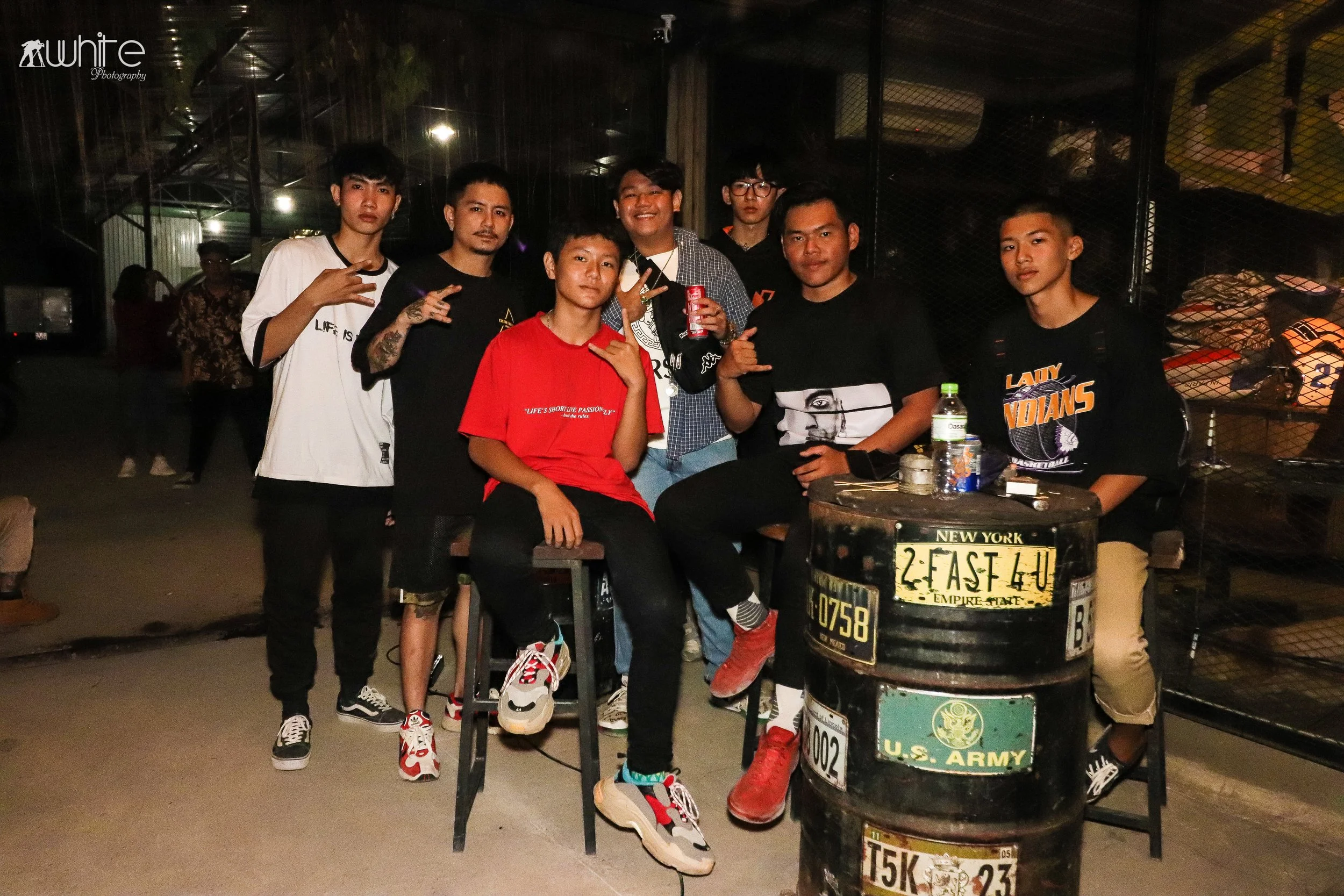 Group of eight young men posing together at night in an urban setting, with some making peace signs or other hand gestures. There are two black barrels with U.S. Army and license plate stickers in the foreground. The background includes a wire fence 