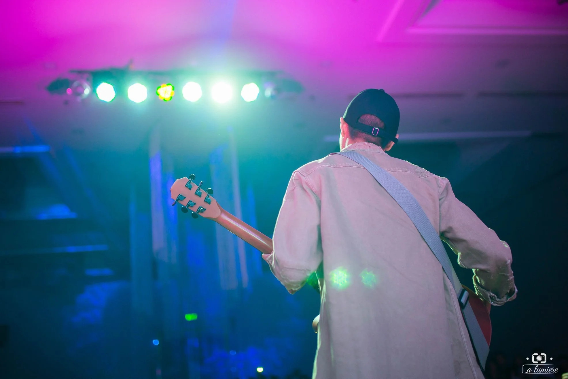 A musician wearing a beige jacket and black cap playing an electric guitar on a stage with bright colored lights in the background.