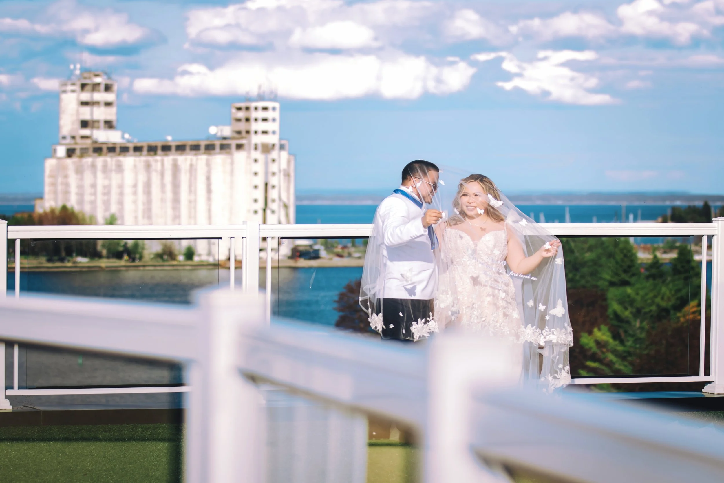 A couple celebrating their wedding on a rooftop terrace, with a cityscape, water, and blue sky in the background. The bride is in a white wedding gown with a veil, and the groom is in a white suit, both smiling and holding a small object.