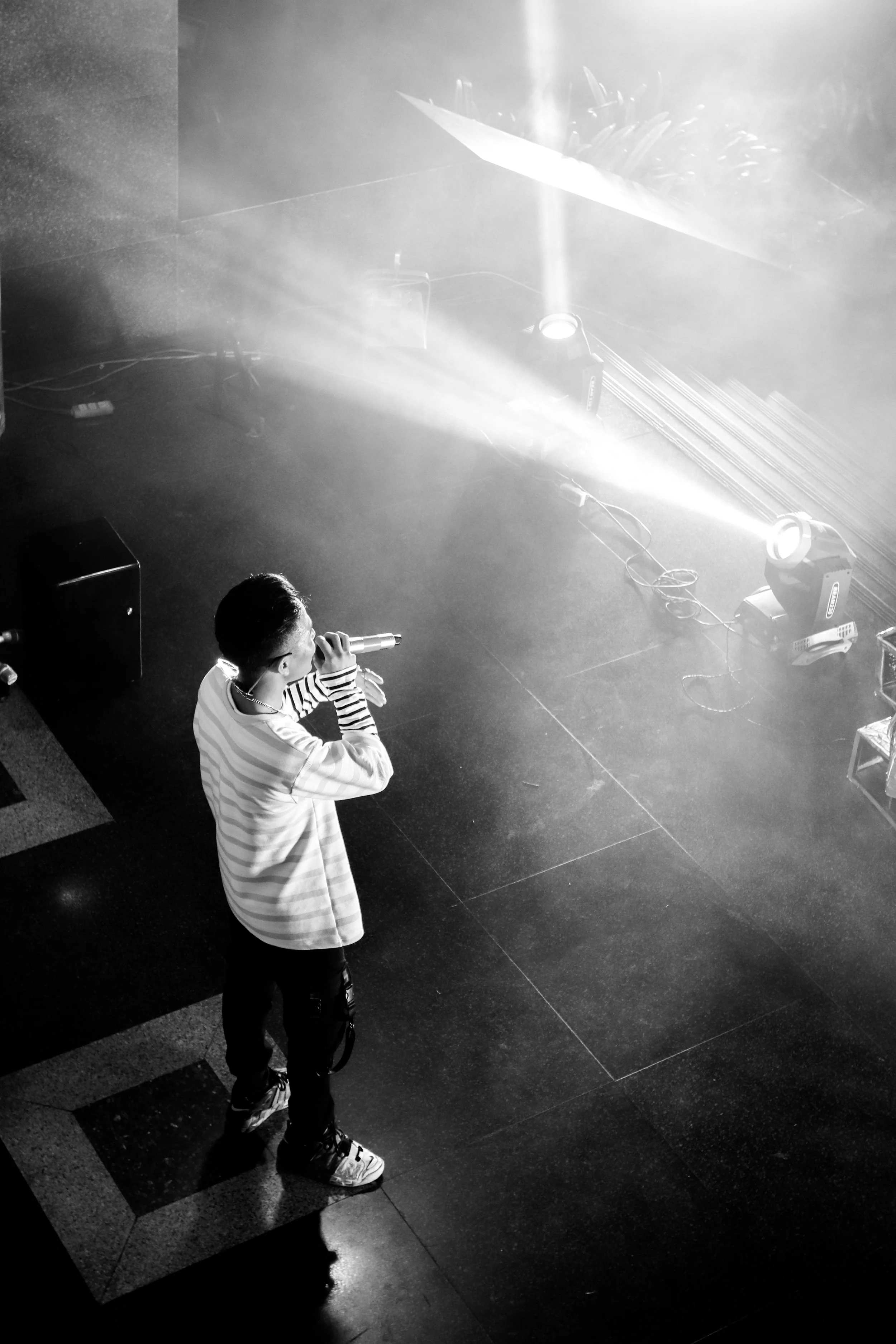 Young person performing on stage, holding a microphone, with stage lights and equipment around, in black and white.