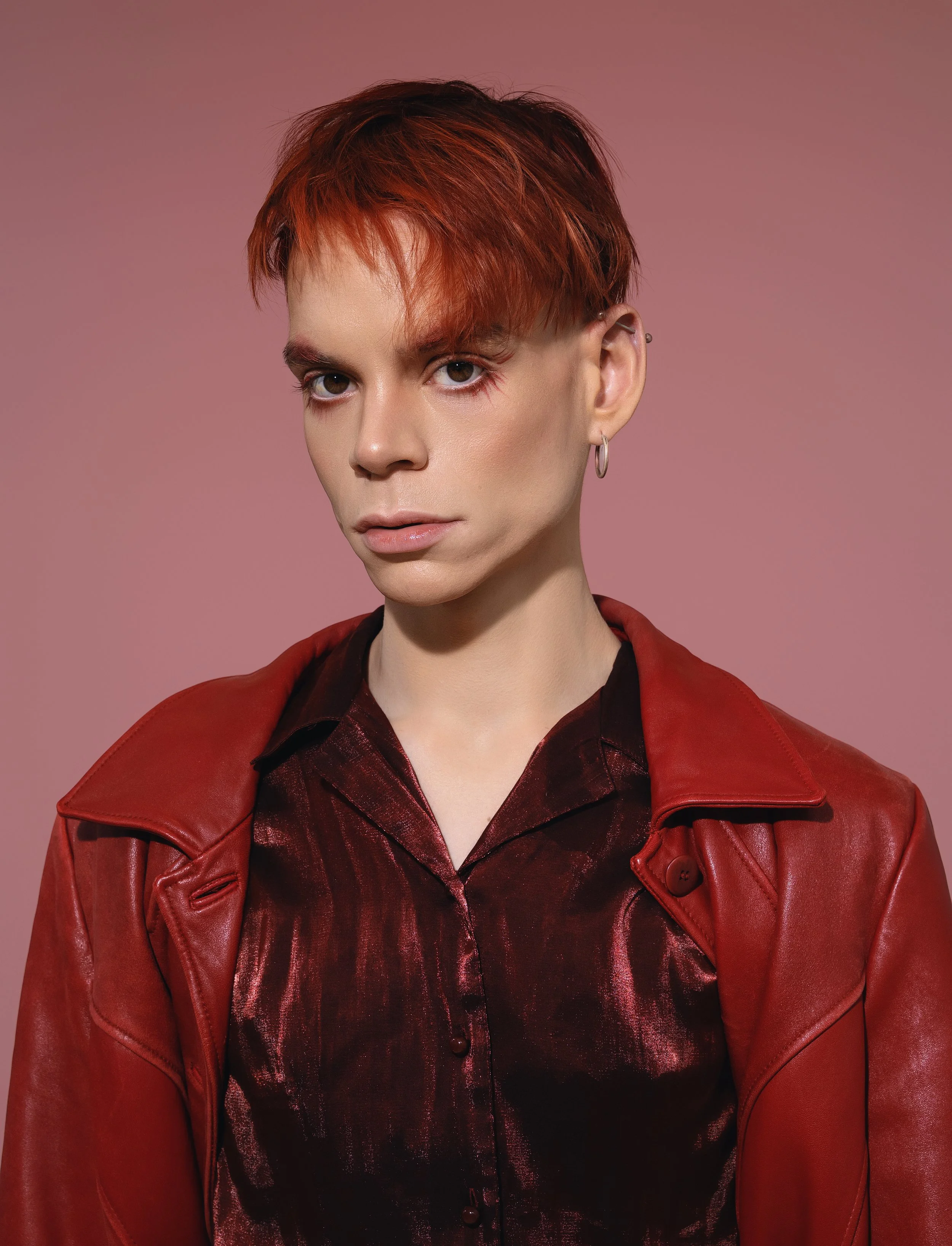 Portrait of a person with short red hair, wearing a red leather jacket and a dark red shirt, against a pink background.