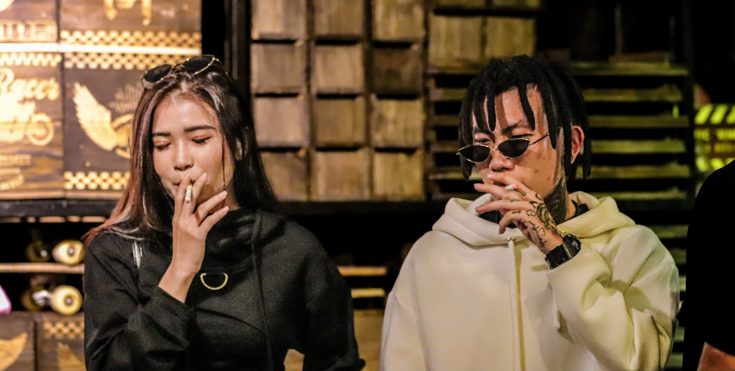 Two young adults, a woman and a man, smoking cigarettes in an indoor setting with wooden panels in the background.