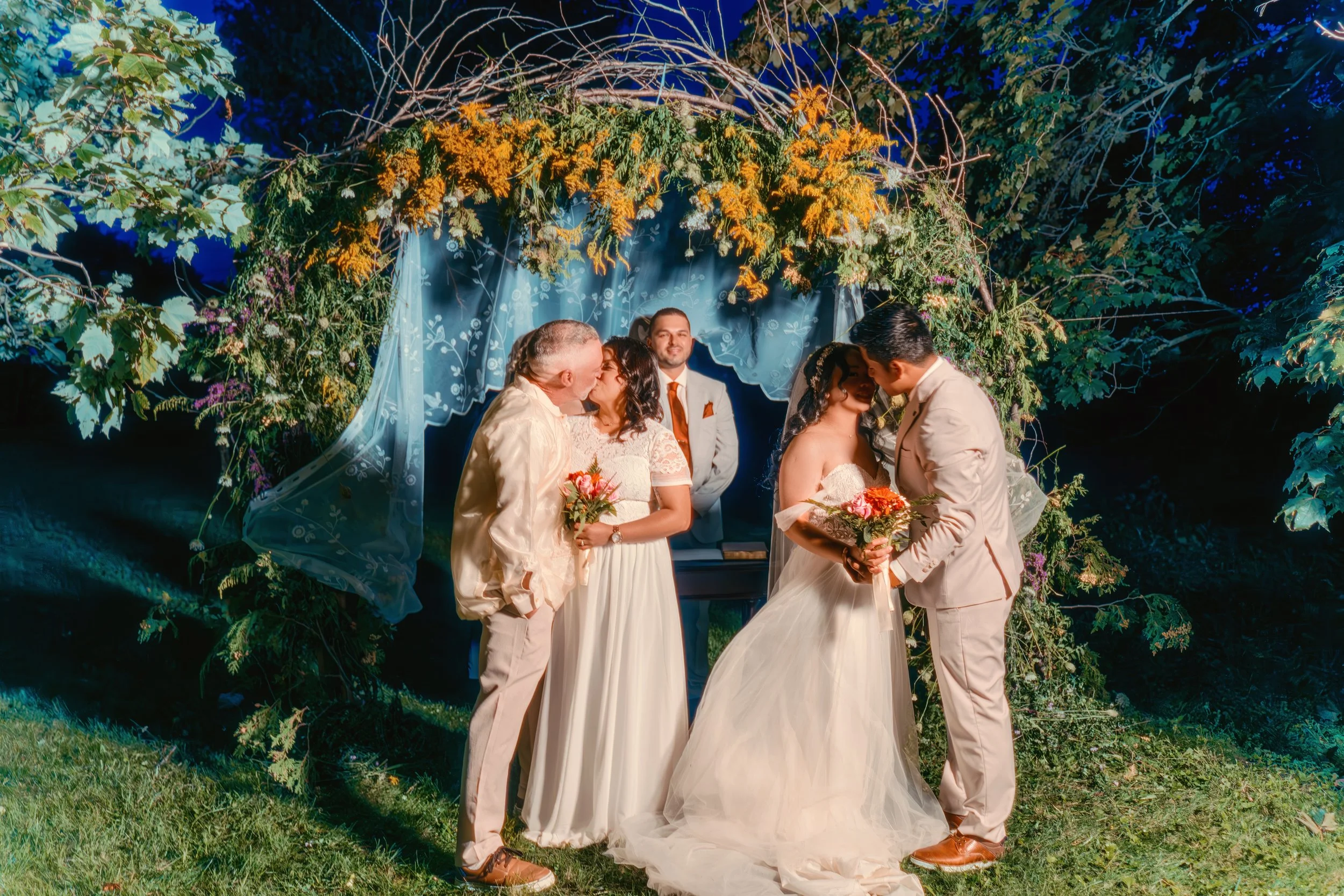 A wedding ceremony outdoors at night with a bride and groom kissing under a floral arch, surrounded by friends and family.