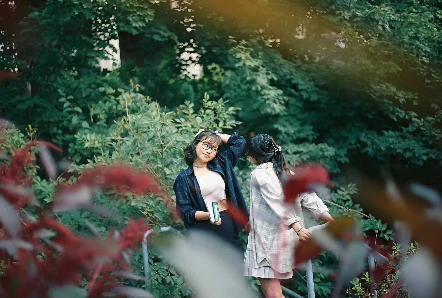 Two young women talking outdoors among green and red foliage, one with glasses holding books, the other with a ponytail, both dressed casually.