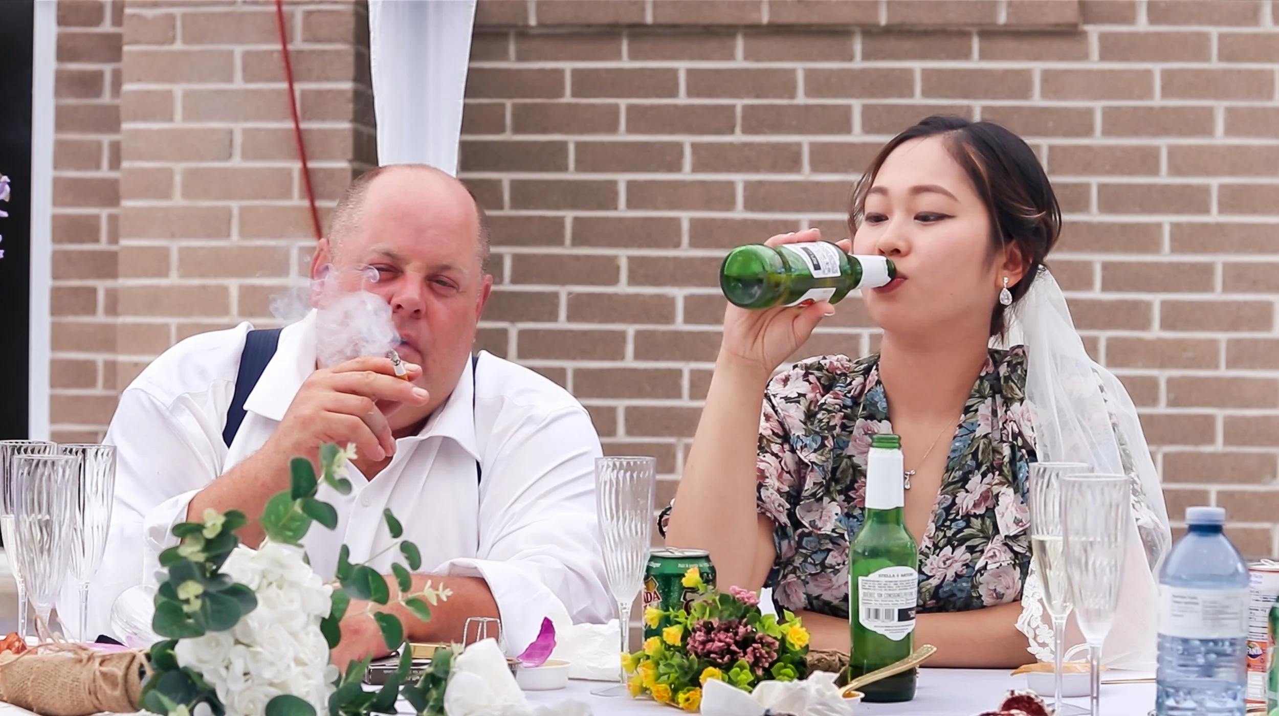 A man and a woman sitting at a dining table, with the man smoking and the woman drinking from a green glass bottle, set against a brick wall.