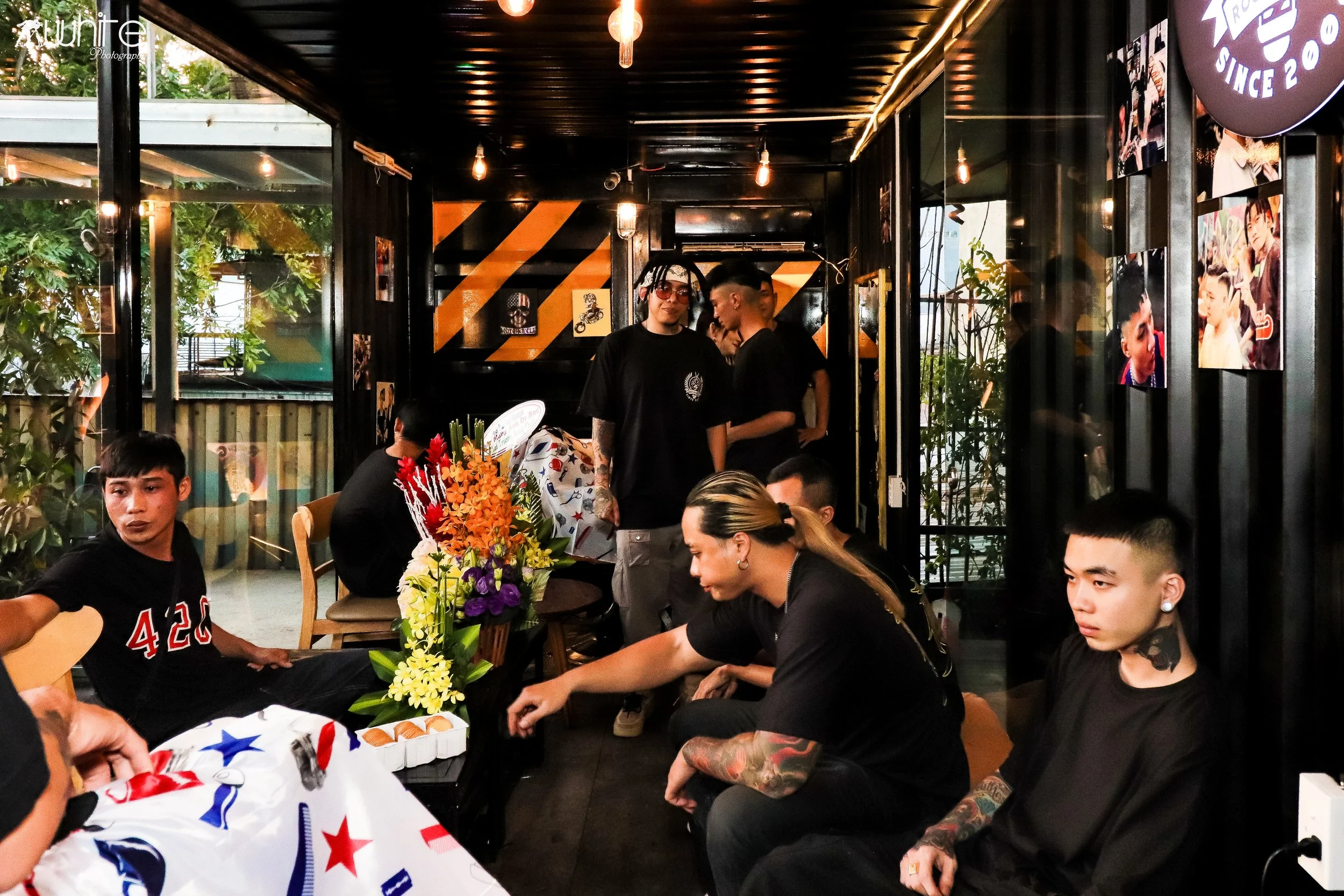 People gathered inside a cozy, decorated room, possibly for a celebration or gathering, with flowers, photos on the wall, and casual attire.