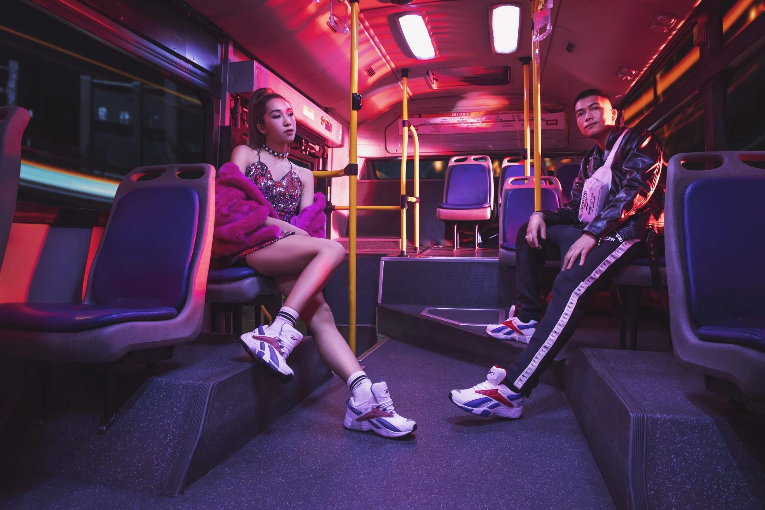 Two young people sitting on a city bus at night, illuminated by pink and purple neon lights. The woman on the left is dressed in a sparkly top, pink fur coat, and white sneakers, with her legs crossed and a pensive expression. The man on the right is