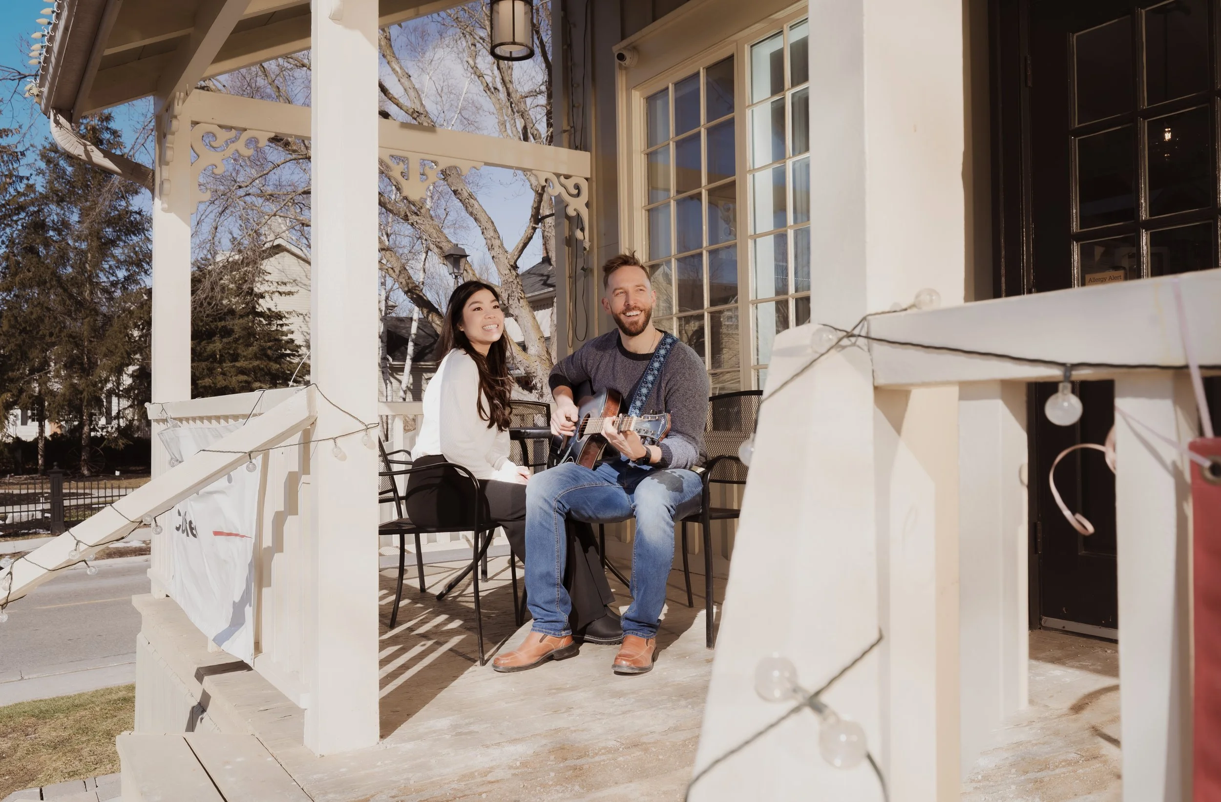 A man and woman sitting on a porch, smiling, with the man playing a guitar. It is daytime with clear skies, and there are trees and houses in the background.