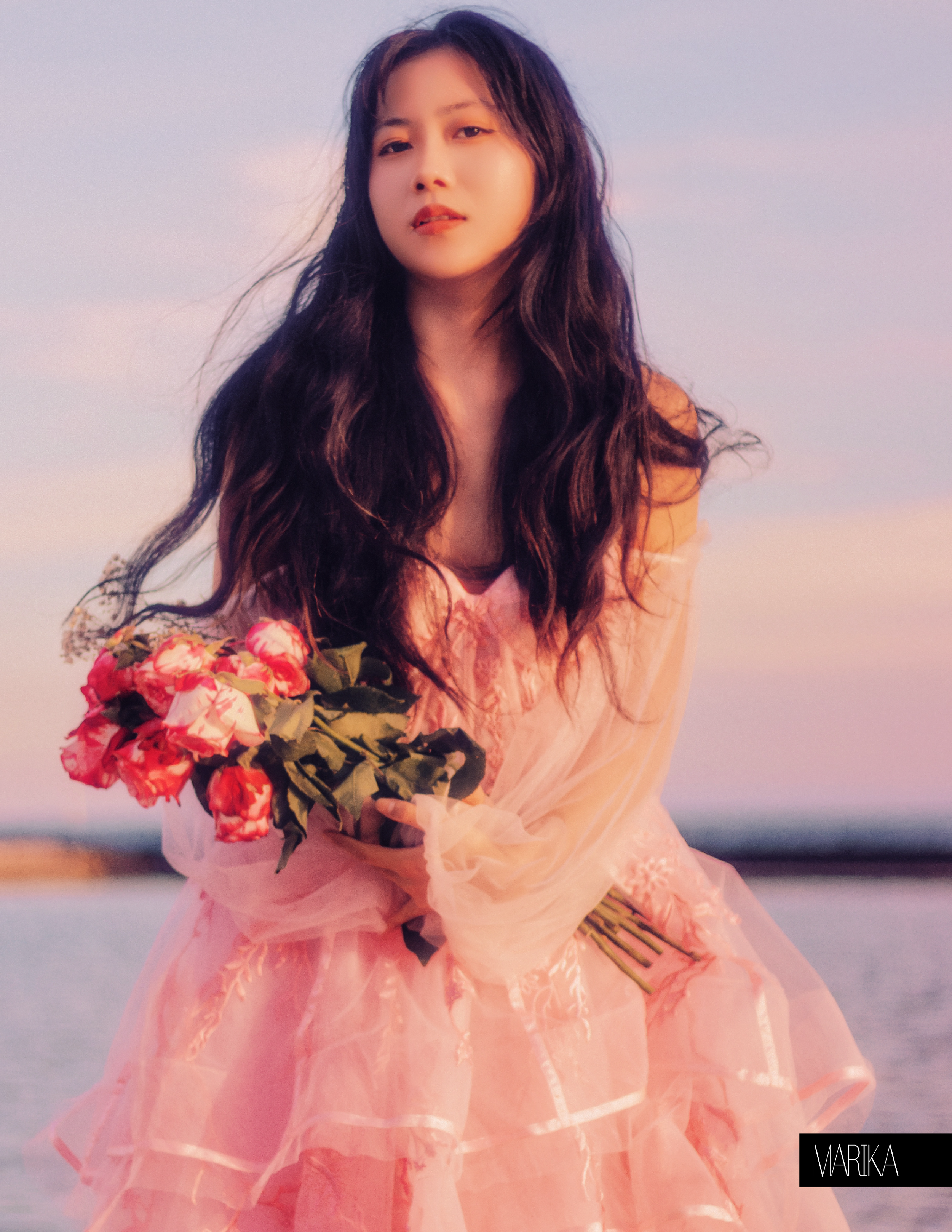 A woman with long, dark hair holding a bouquet of pink and white flowers, wearing a flowing pink dress outdoors during sunset.